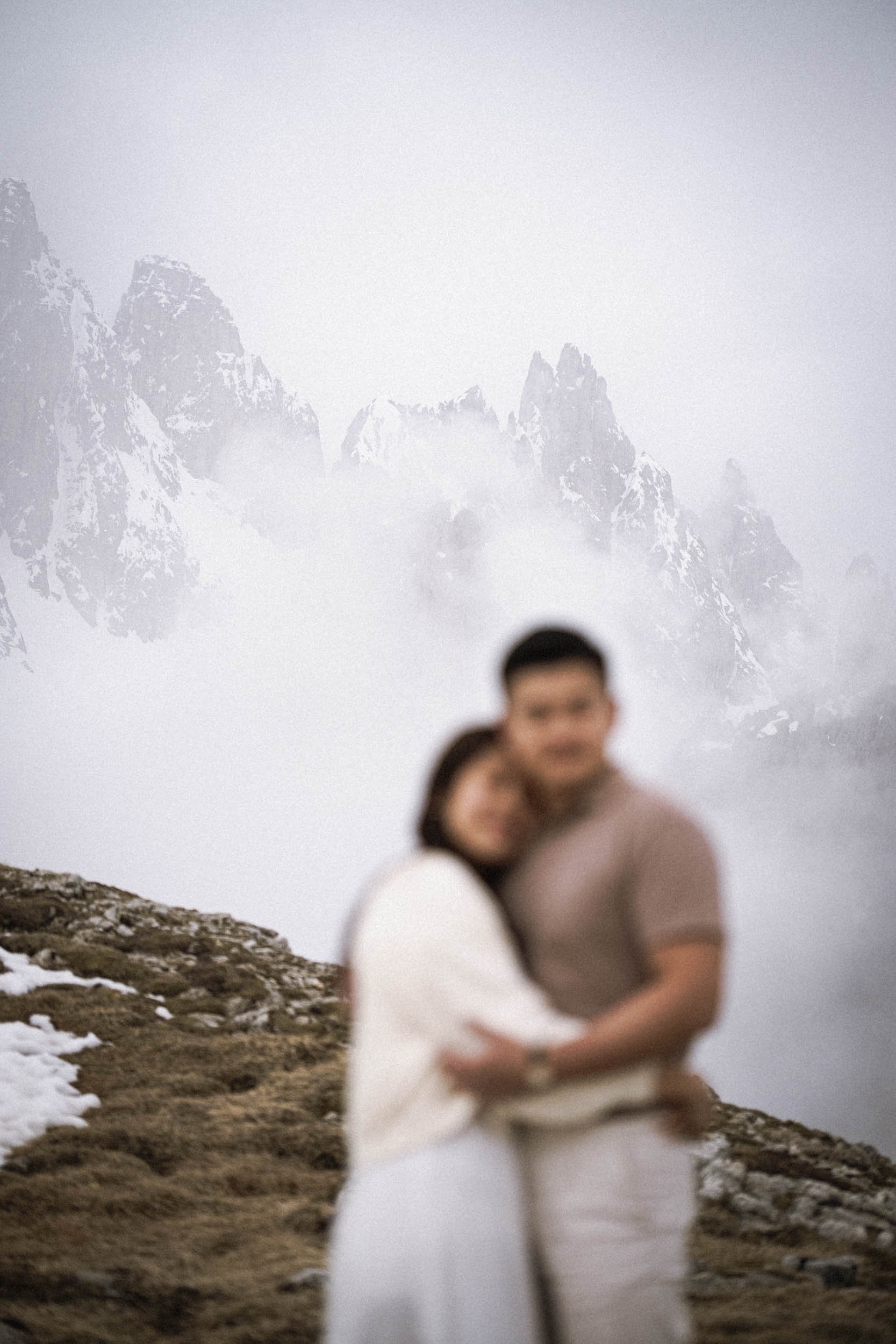 Ein unscharfes Foto eines Paares, das sich umarmt, vor einer Berglandschaft mit schneebedeckten Gipfeln und Nebel.