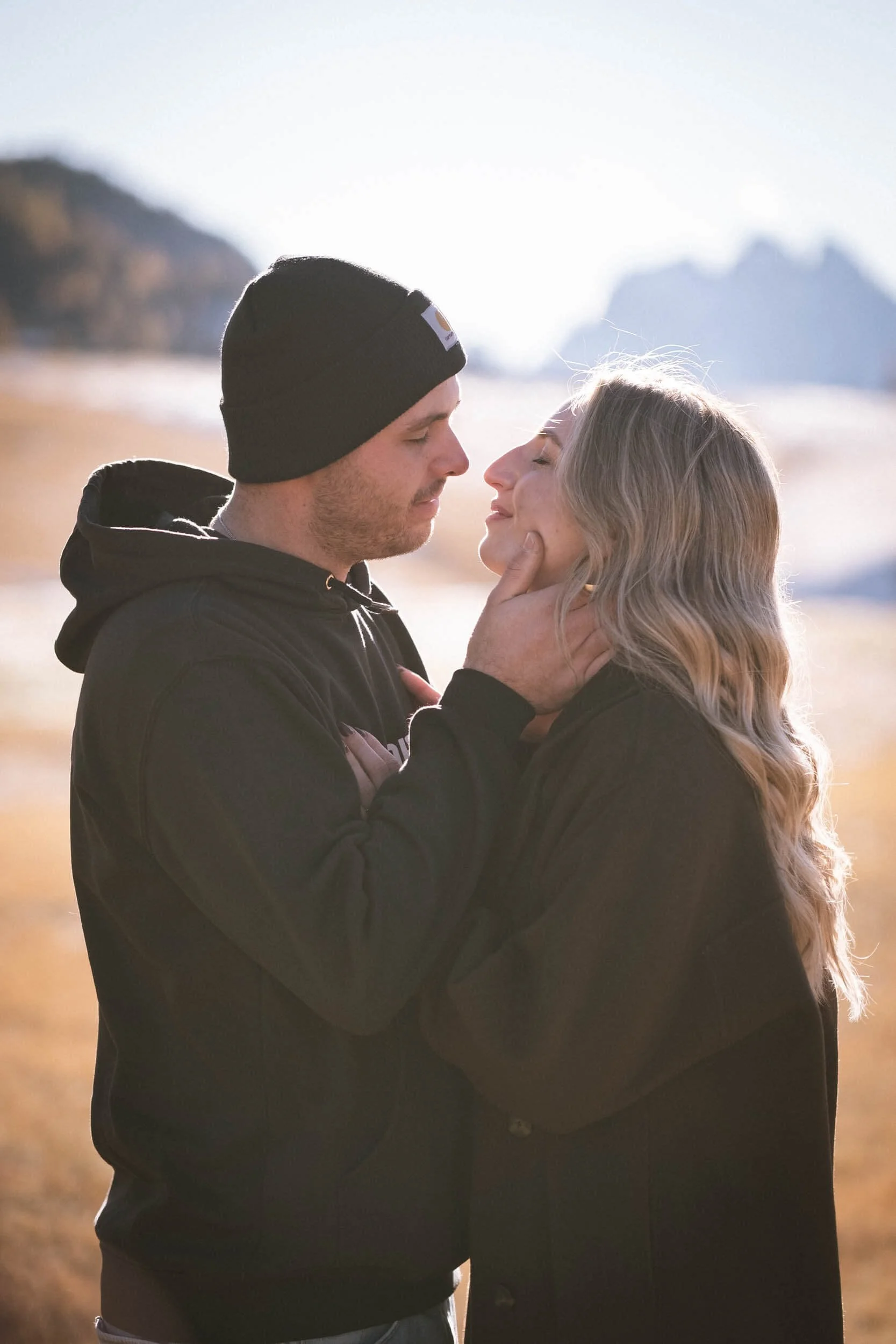 Ein Mann und eine Frau stehen sich nahe am Strand gegenüber, der Mann hält die Frau zärtlich im Gesicht, während die Frau lächelnd ihre Augen schließt.