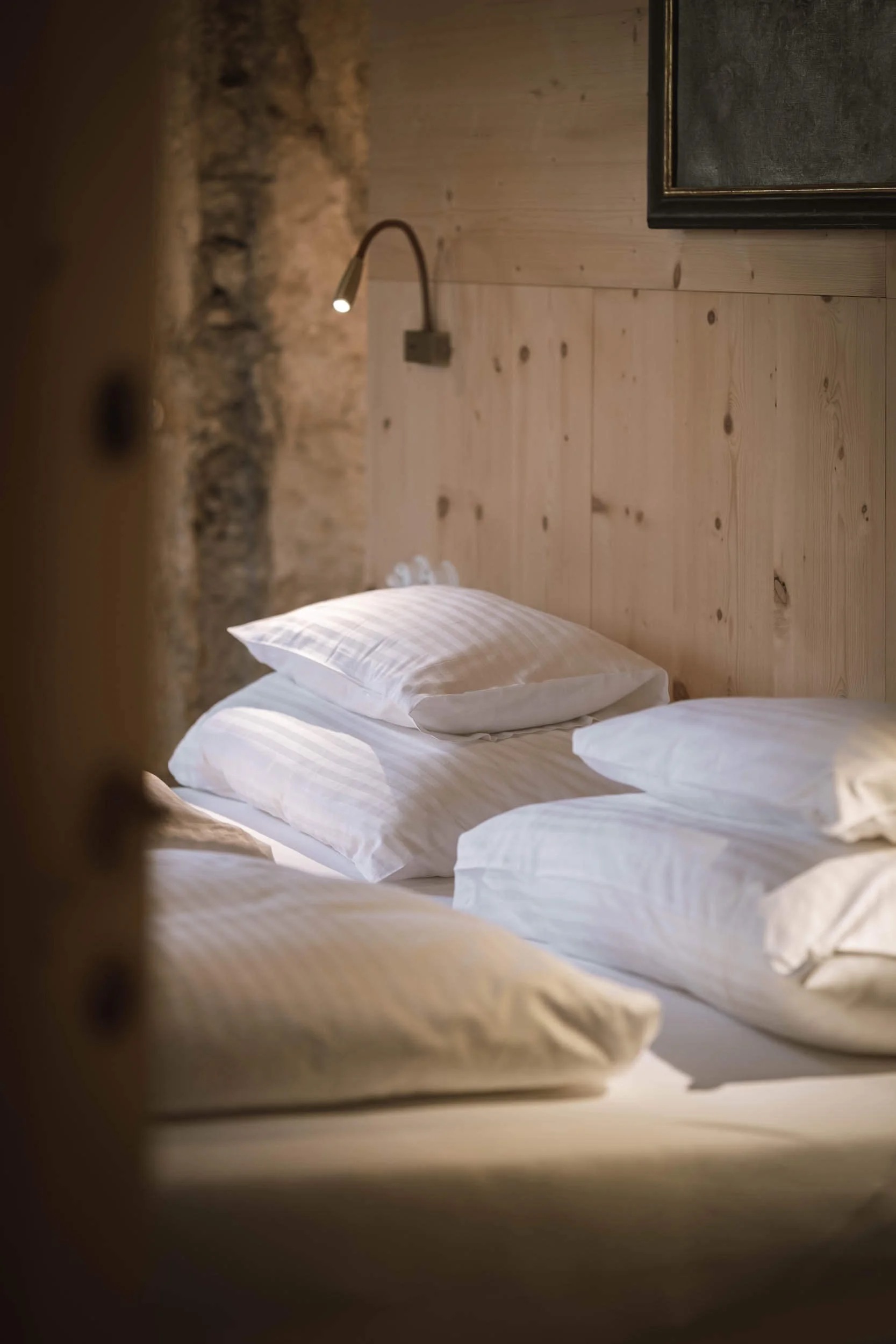 Bett mit weißen Kissen und Bettwäsche in einem rustikalen Schlafzimmer mit Holz- und Steinwänden, beleuchtet von einer Ganzkopf-Wandlampe.