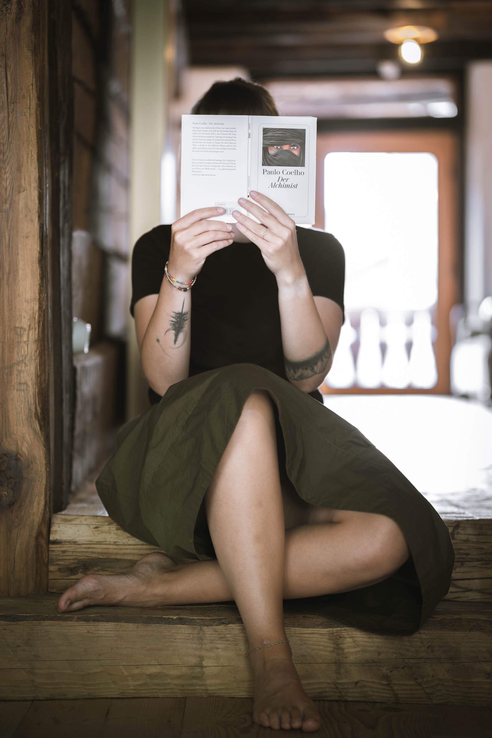 Eine Person sitzt in einem Holzhaus, liest ein Buch und hat Tattoos am Arm.
