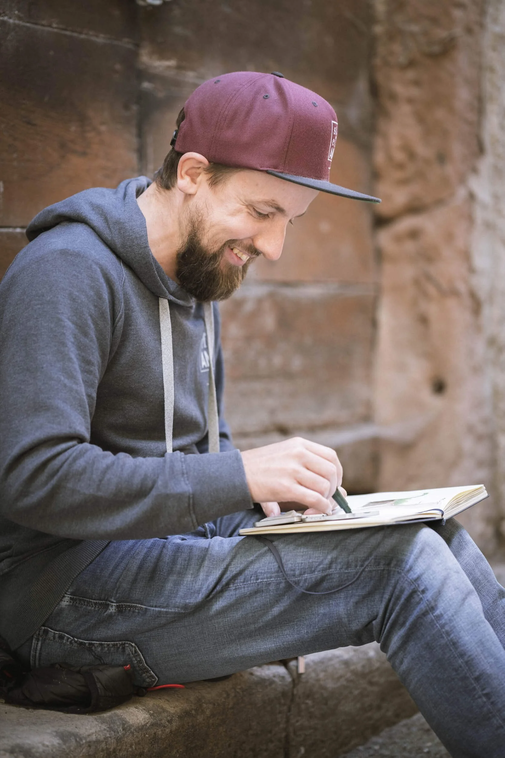 Ein lächelnder Mann mit Bart sitzt auf einer Steinbank, trägt eine graue Kapuzenjacke und eine dunkelrote Mütze, und liest in einem offenen Buch.