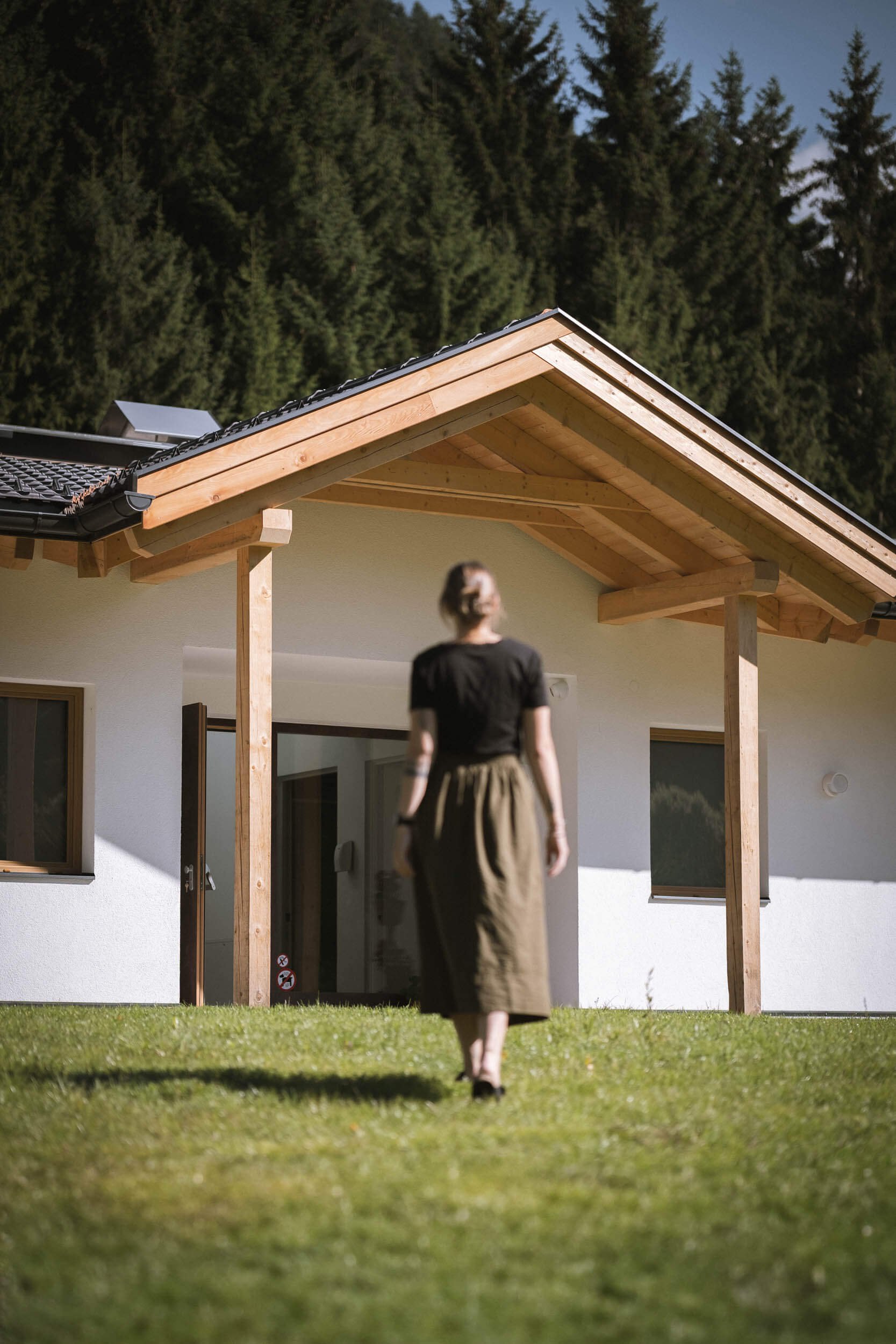 Frau läuft auf grünem Rasen in Richtung eines modernen Hauses mit weißer Fassade und Holzdachüberstand. Hintergrund mit Wald aus Tannenbäumen.