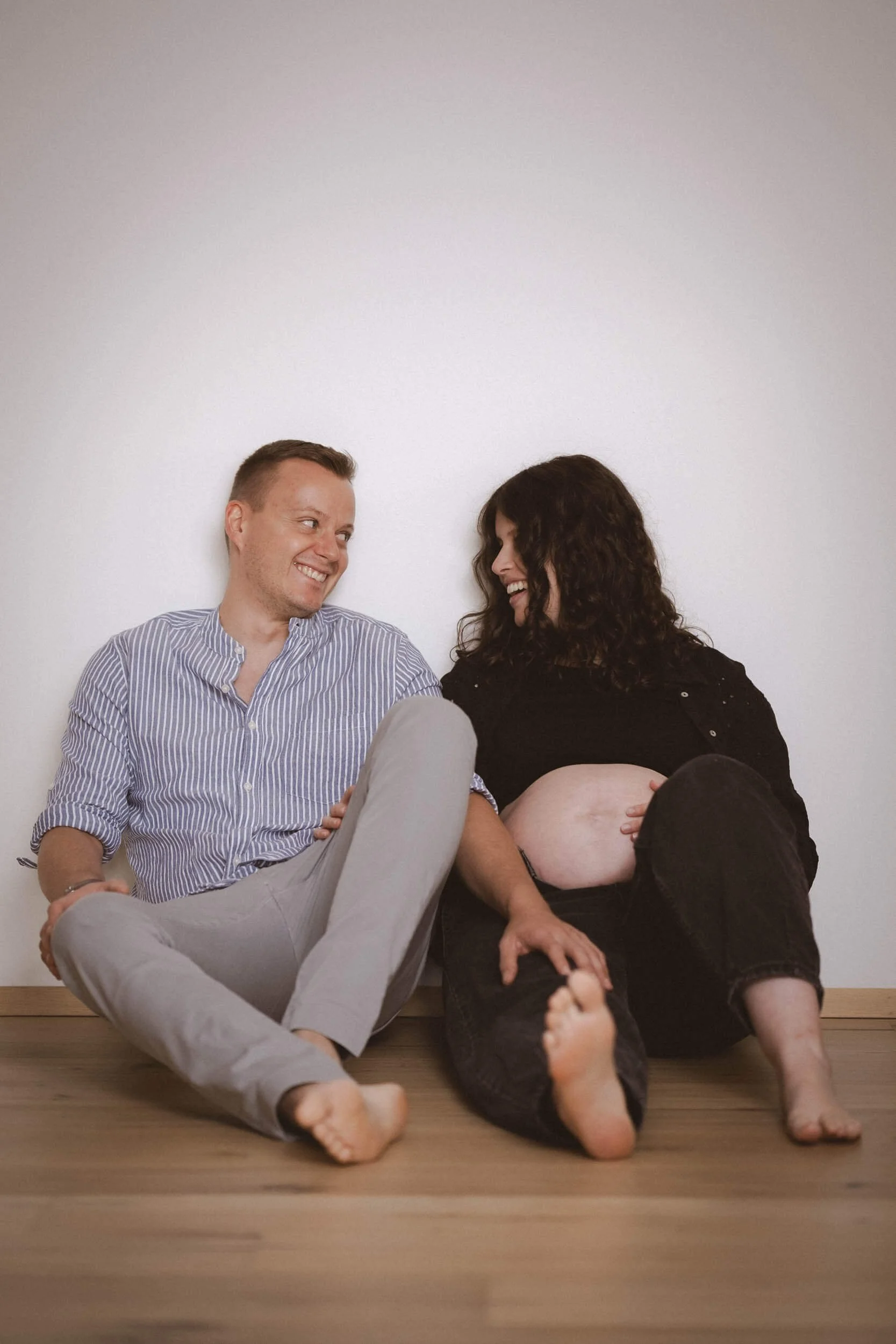Ein glückliches Paar sitzt auf dem Boden. Die Frau ist schwanger, trägt eine schwarze Jacke und eine schwarze Hose, während der Mann ein gestreiftes Hemd und helle Hosen trägt. Sie lachen und schauen sich an, die Frau hat den Bauch gestreichelt.