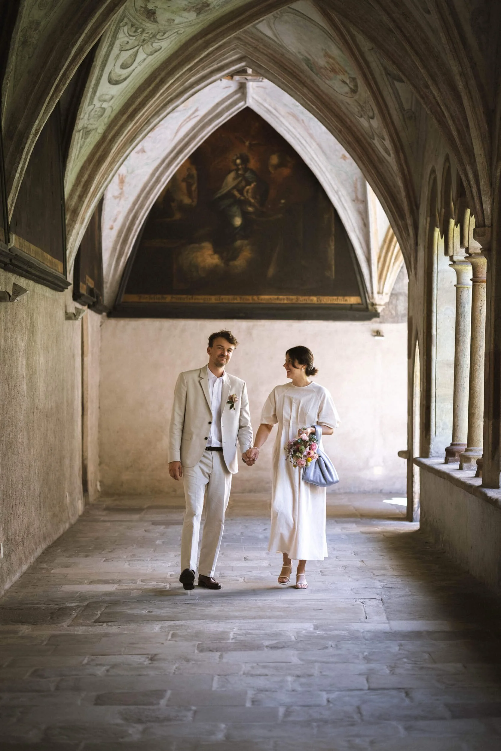 Ein Paar, vermutlich Braut und Bräutigam, hält Händchen in einer alten, gotischen Kirche mit einem Gemälde im Hintergrund. Sie trägt weiße Kleidung, sie eine weiße Robe mit Blumenstrauß, er einen hellen Anzug.