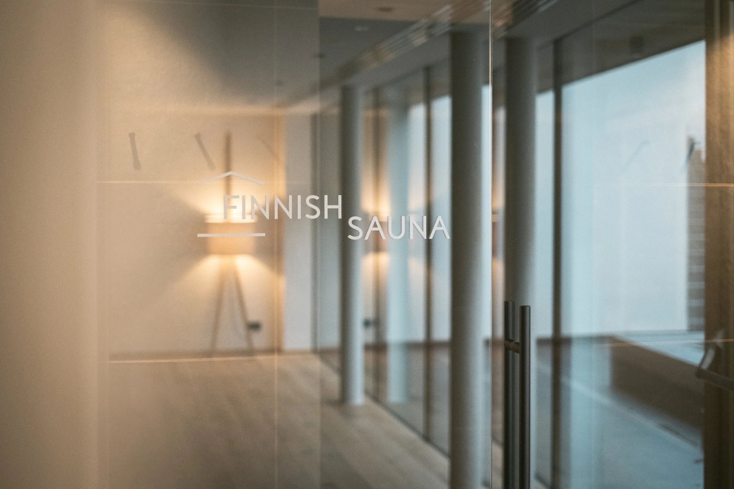 Durchsichtige Tür mit der Aufschrift 'Finnish Sauna' im Inneren eines modernen Spa-Bereichs, mit Holzboden und Lampen an der Wand.