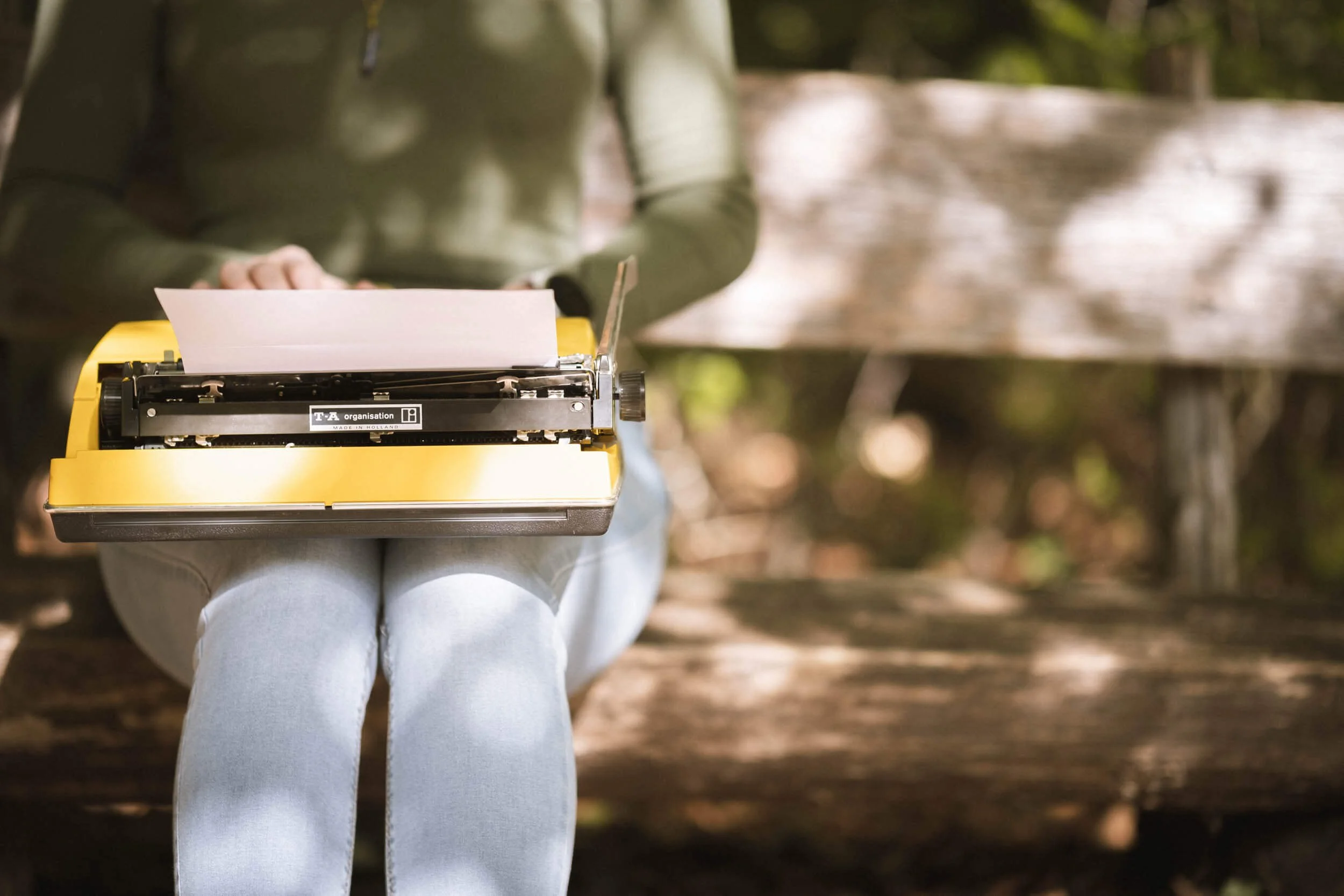Person sitzt auf einer Bank im Freien und tippt auf einer alten, gelben Schreibmaschine.