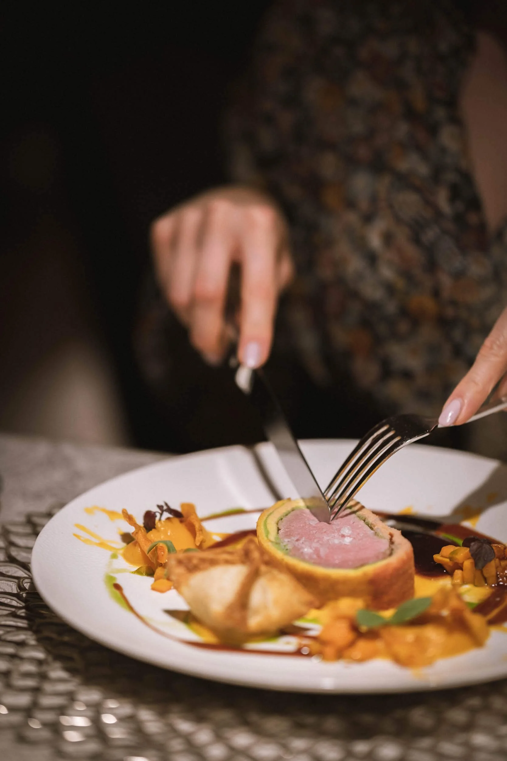 Perspektive auf einen Teller mit kunstvoll angerichtetem Gericht, jemand schneidet mit Messer und Gabel in das Gericht.