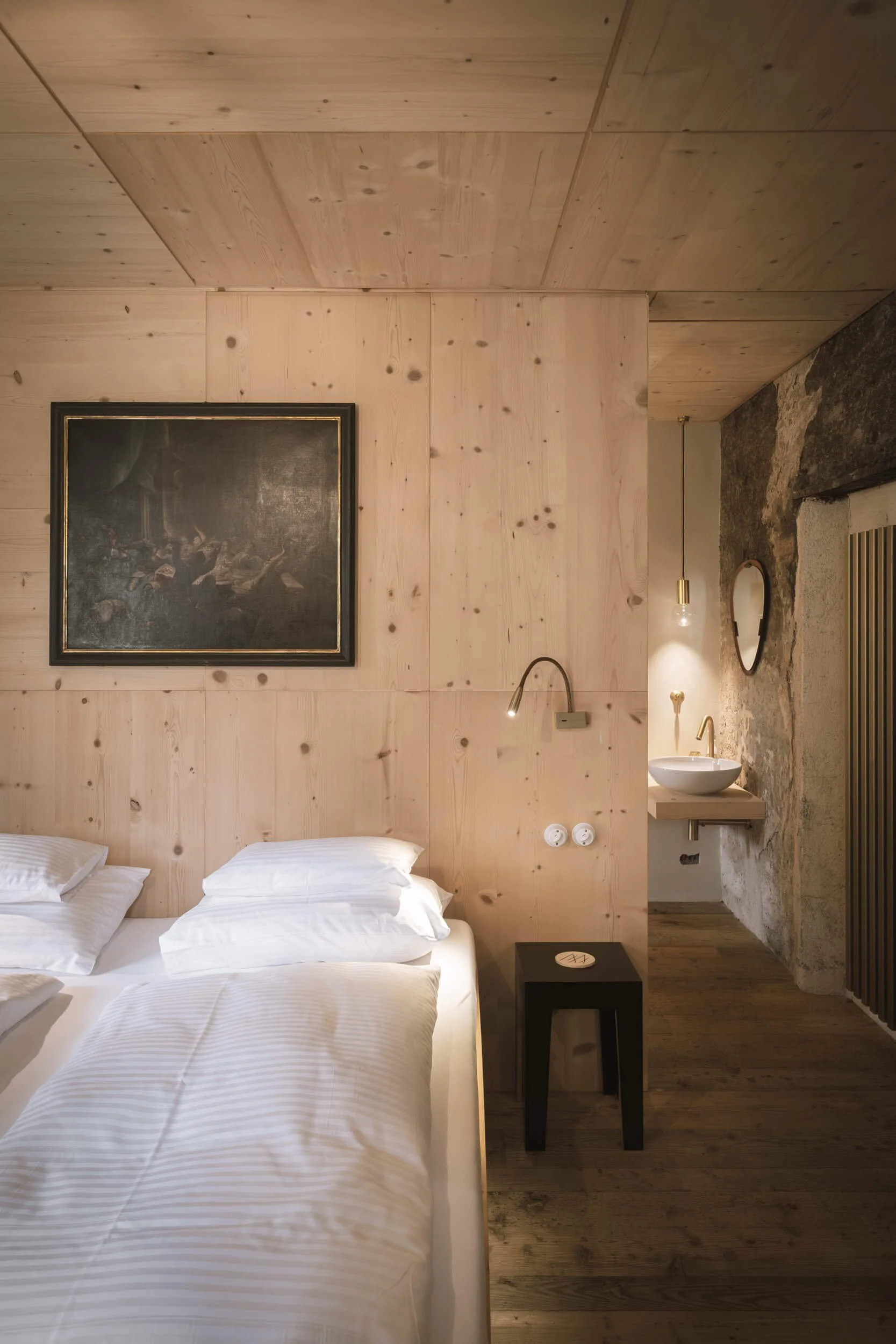 Ein Schlafzimmer mit Holzverkleidung an Wand und Decke, ein Gemälde, ein Nachttisch, eine Bett, ein Wandlampen, ein Waschbecken an einem Felsen, eine kleine runde Spiegel