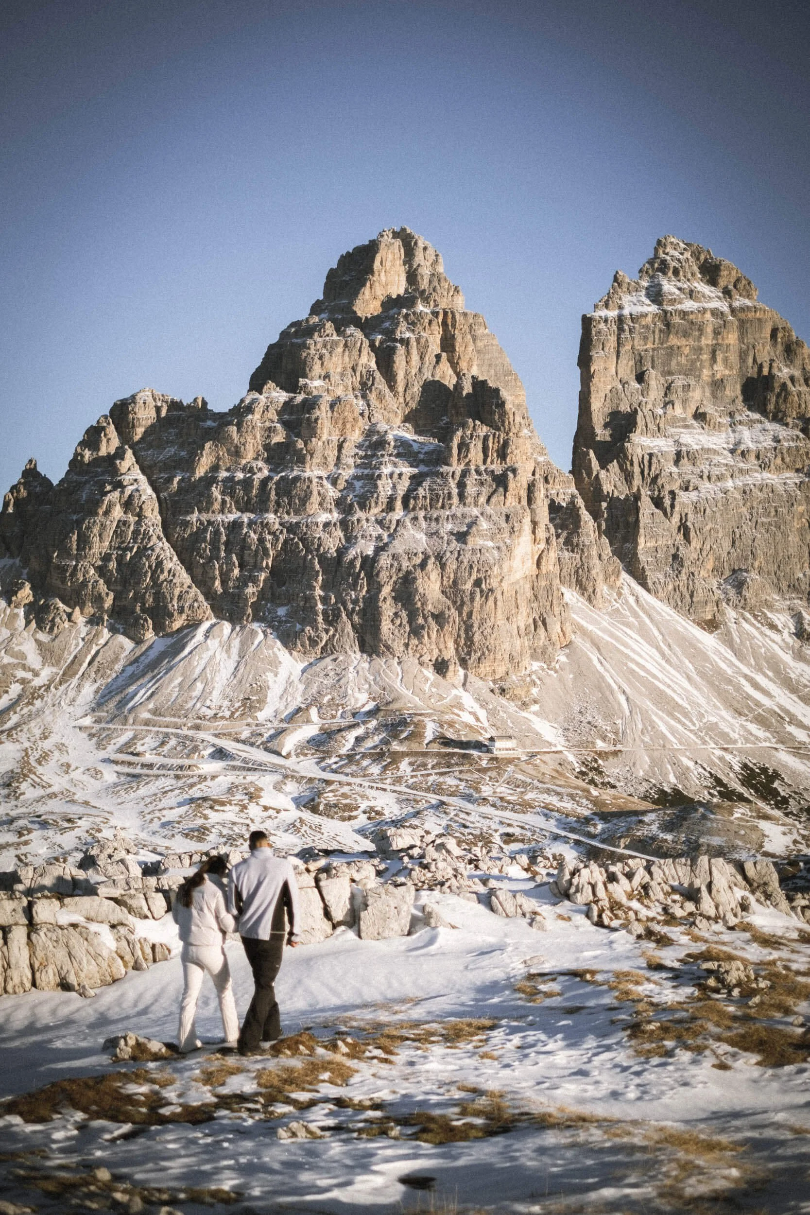 Zwei Menschen, ein Mann und eine Frau, gehen in Schnee vor einer Bergkulisse mit hohen Felsformationen.
