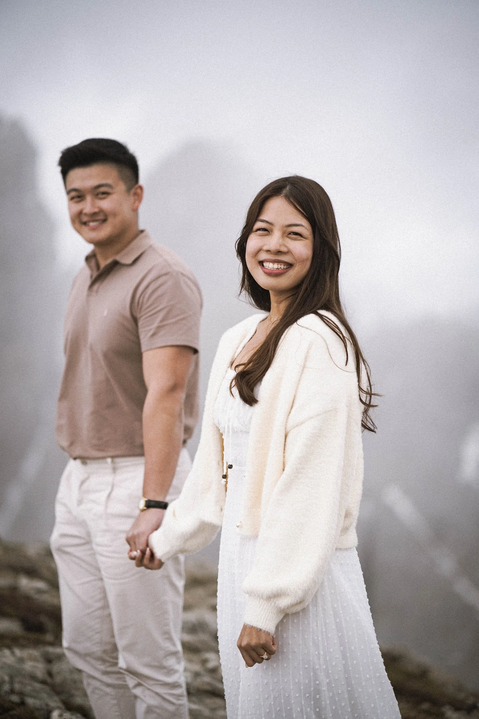 Ein Mann und eine Frau halten Händchen auf einem nebligen Berggipfel, lächelnd, in hellem, casual Kleidung.