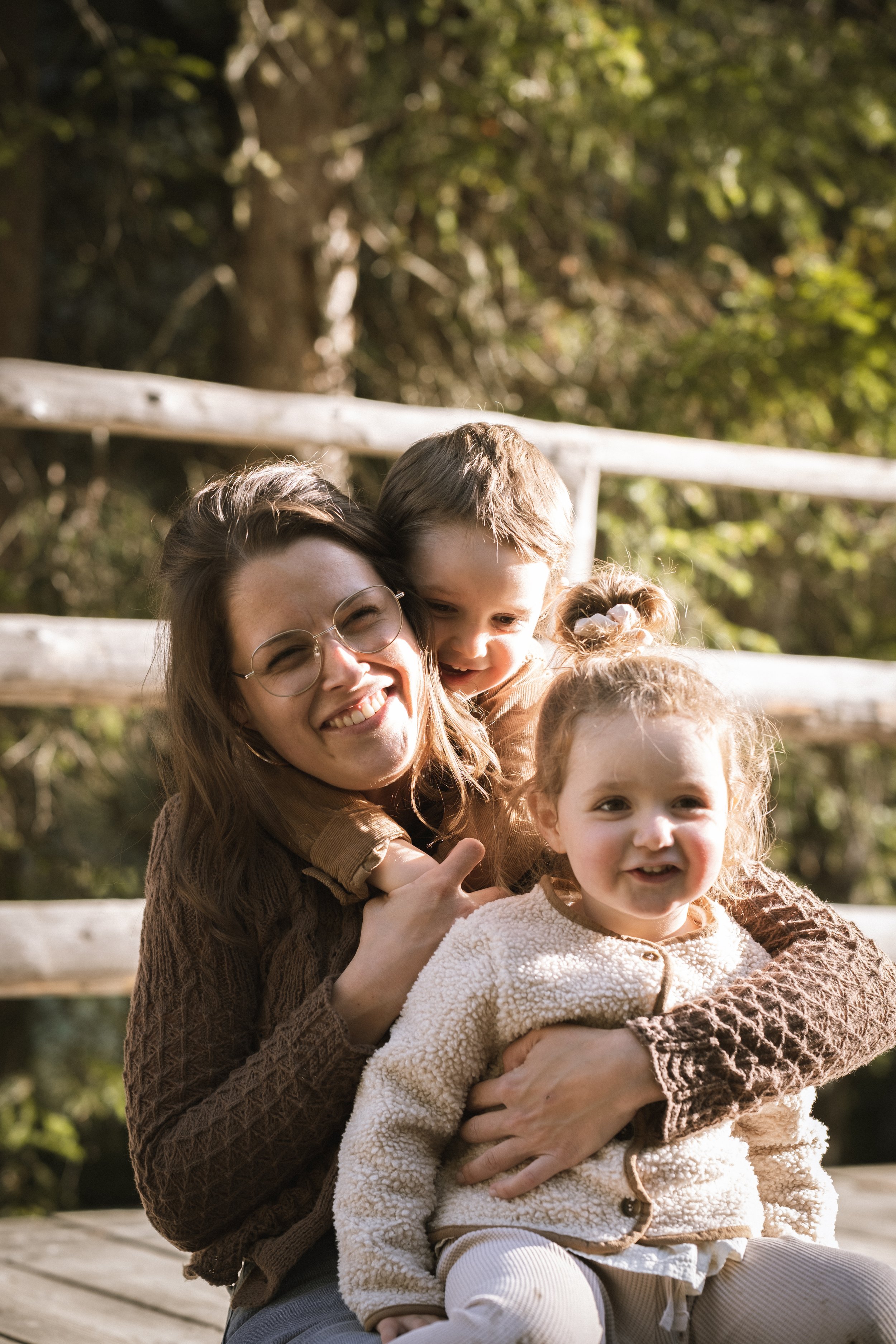 Frau mit zwei kleinen Mädchen beim Umarmung im Freien, lächelnd, auf Holzbrücke, im Hintergrund Bäume.