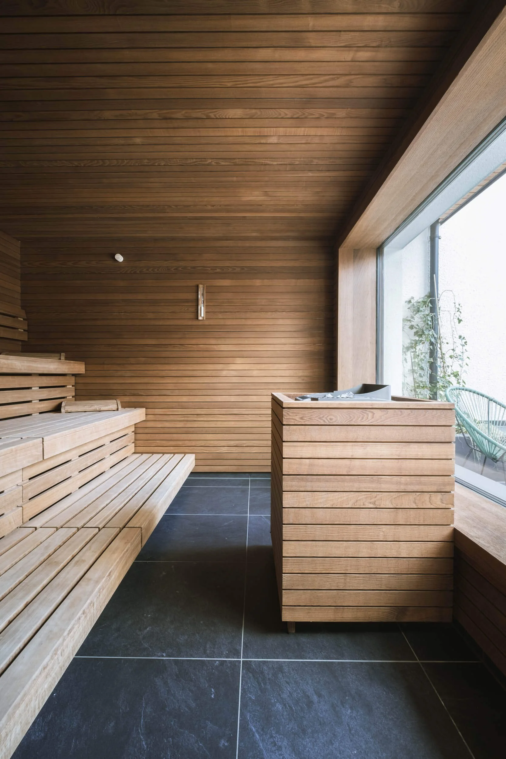 Innenraum einer Sauna mit Holzverkleidung, dunklen Fliesenboden und großen Fenstern, die einen Blick nach draußen auf einen Sitzbereich mit grünen Stühlen bieten.