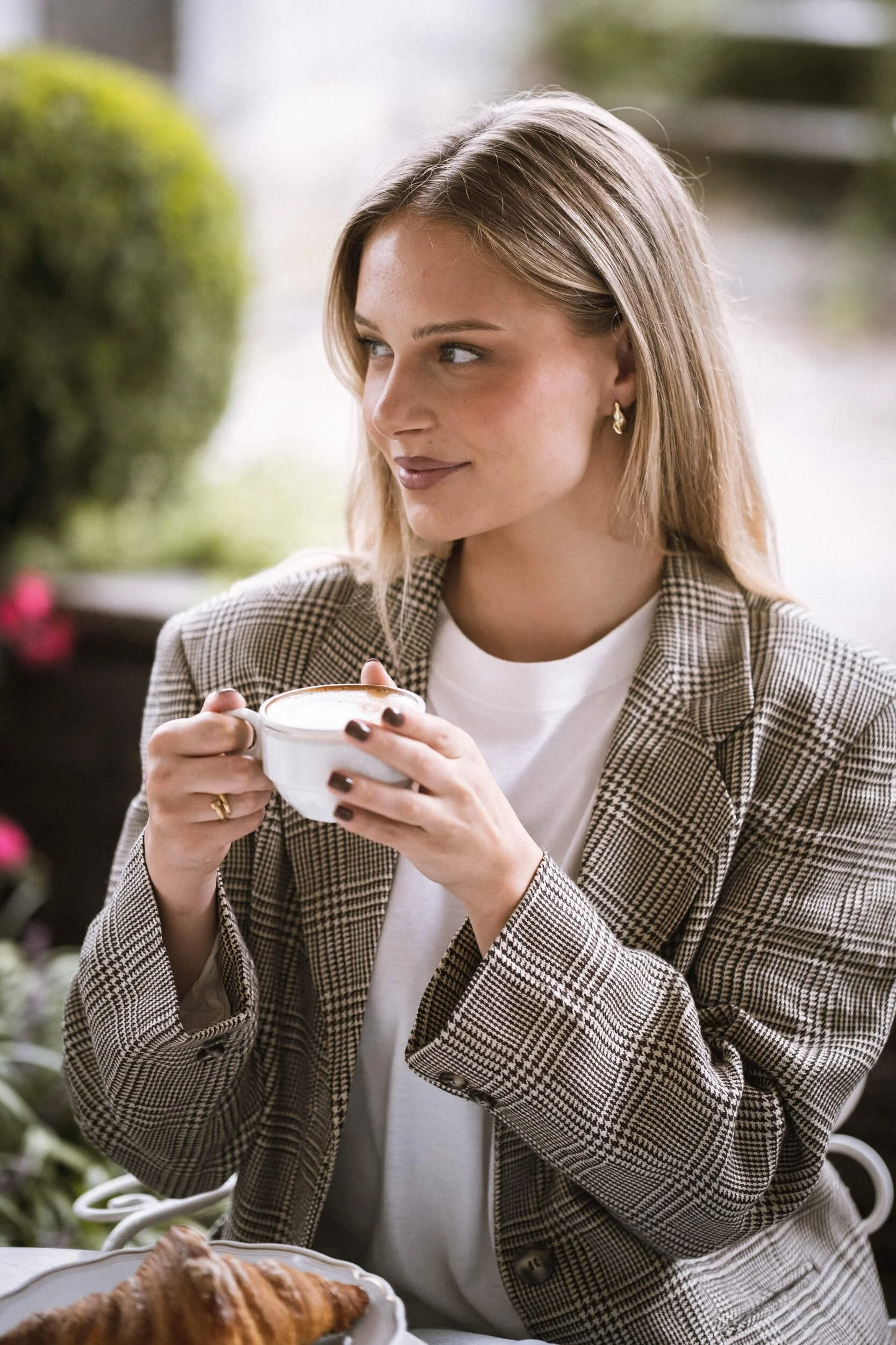 Frau mit blonden Haaren trägt einen karierten Blazer und trinkt einen Kaffee in einer Tasse, im Vordergrund liegt ein Croissant auf einem Teller.