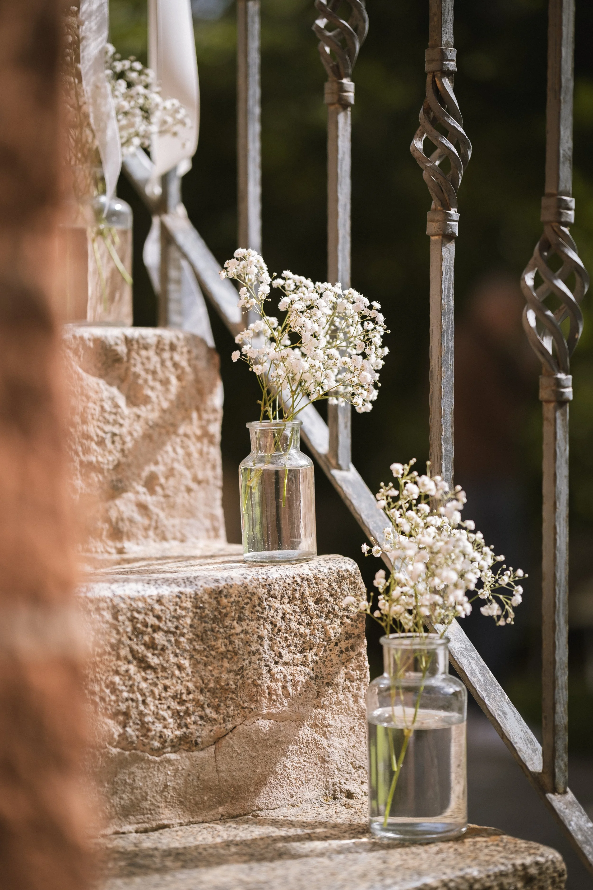 Zwei Gläser mit weißen Blumen auf Steinstufen neben einer Eisenleitplanke bei Sonnenlicht.