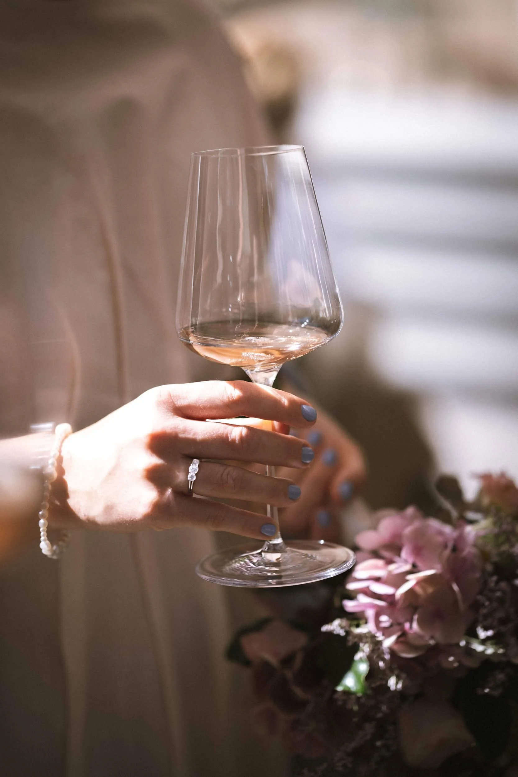 Eine Person hält ein Weinglas mit roséfarbenem Wein. Die Person trägt ein Onyx-Ring und eine Perlenarmband. Im Hintergrund sind Blumen sichtbar.