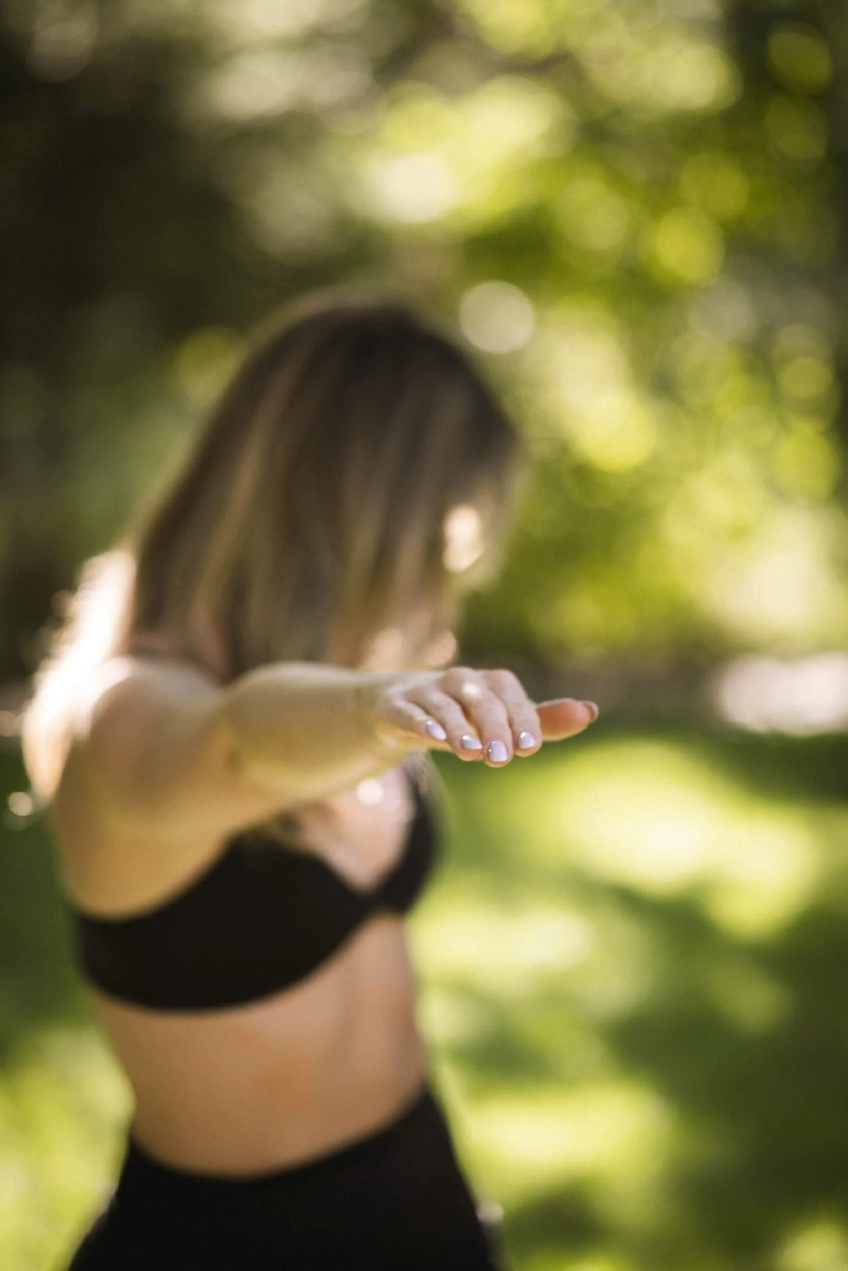 Frau in schwarzem Sport-BH macht Yoga-Übung im Park, mit Fokus auf ausgestreckte Hand. Hintergrund mit Sonnenlicht und grünen Blättern.