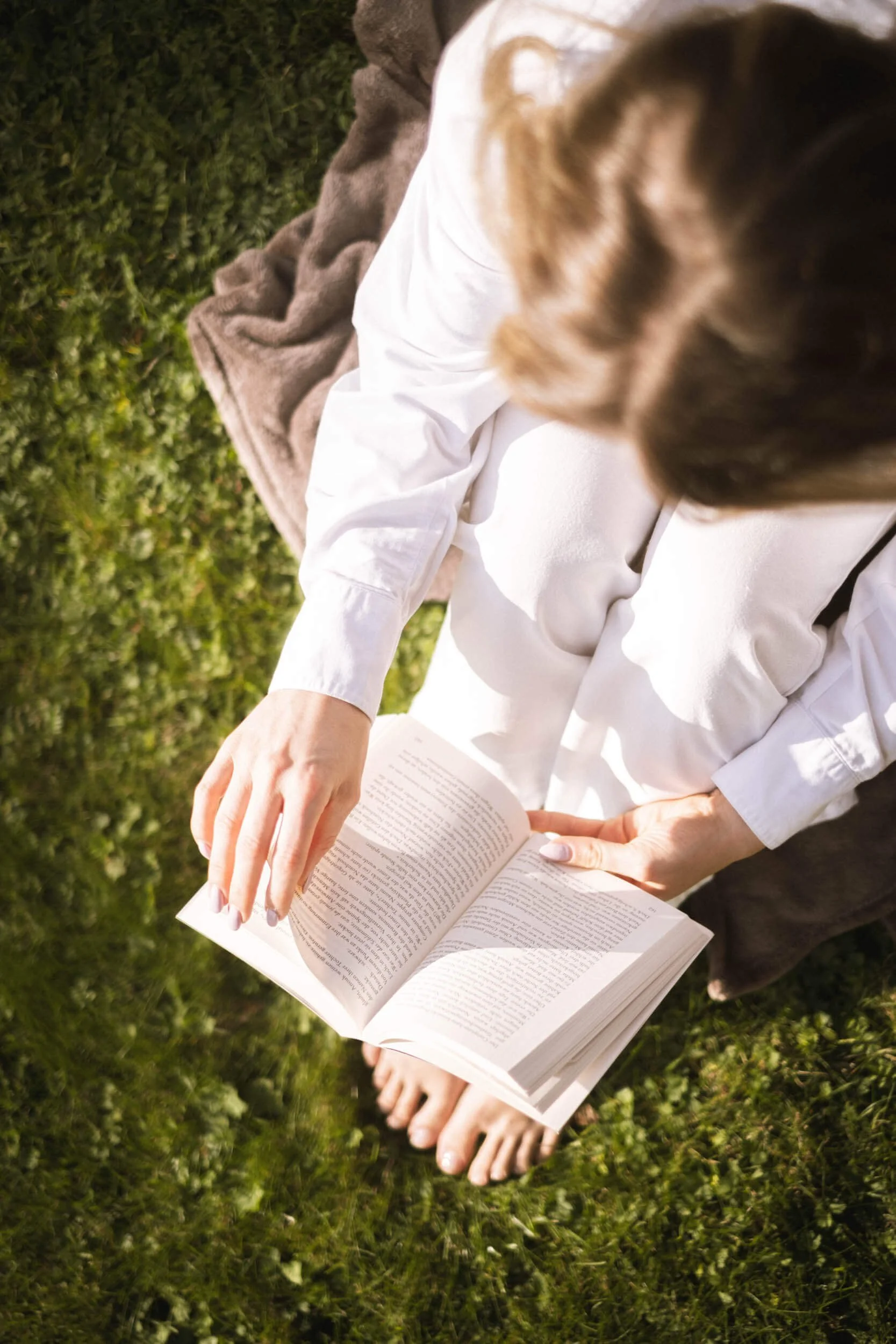 Eine Person sitzt barfuß auf grünem Gras, liest ein Buch. Im Hintergrund liegt eine braune Decke.