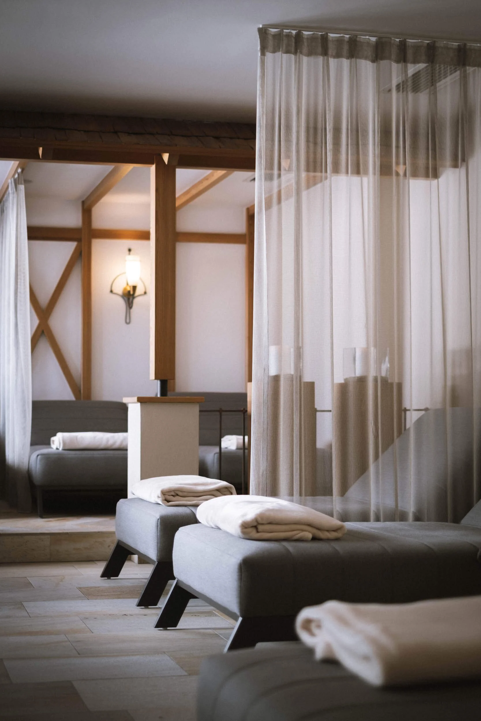 Ein Spa- oder Wellnessbereich mit mehreren Liegen, die mit weißen Handtüchern bedeckt sind, und einem durchsichtigen Vorhang, der einen Bereich abtrennt. Im Hintergrund sind Holzelemente und eine Wandlampe sichtbar.