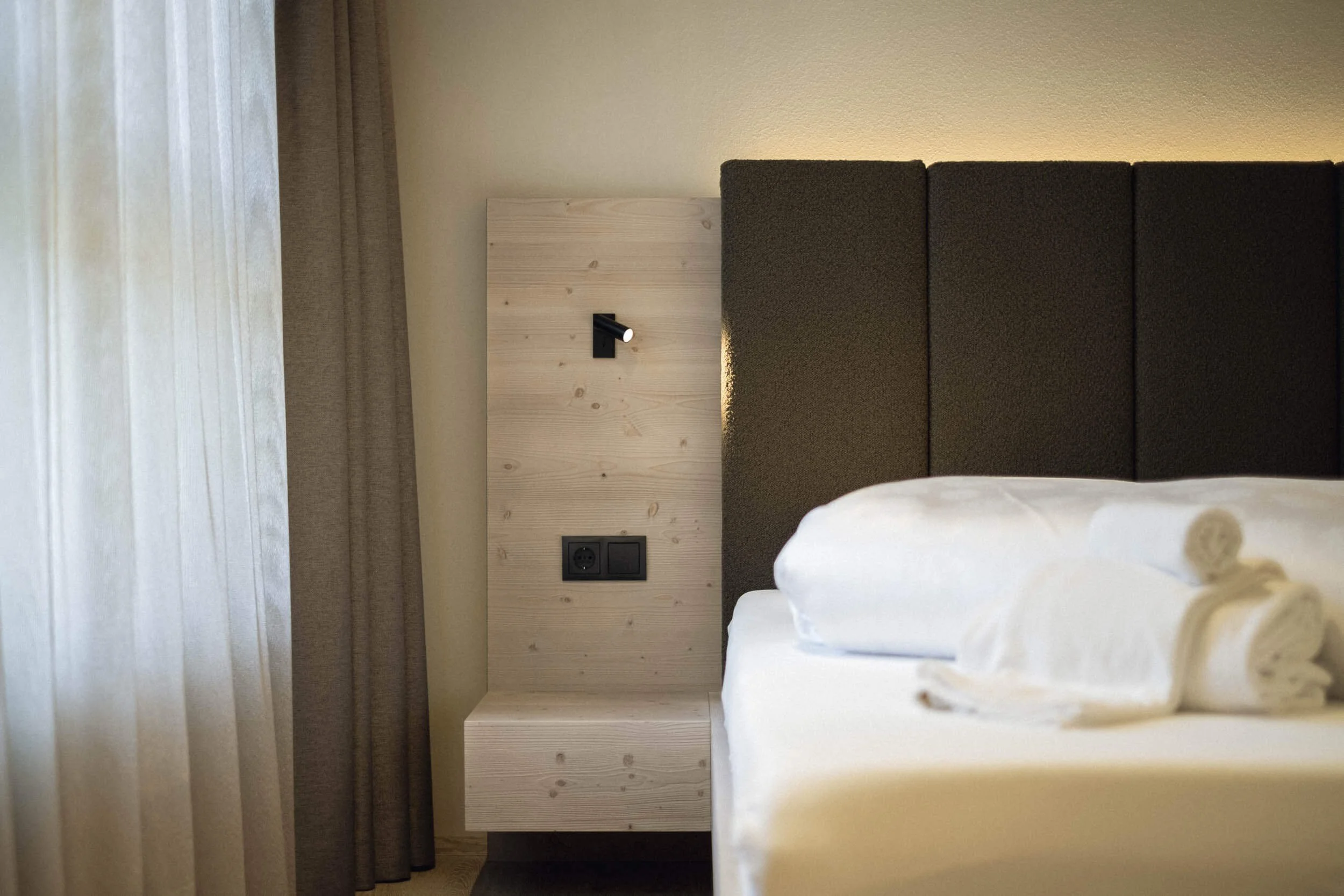 Ein Hotelzimmer mit einem Bettrahmen, einem Kopfteil und einem Laken, ein Nachttisch mit einer Wandlampe und Steckdosen, Vorhänge vor einem Fenster.