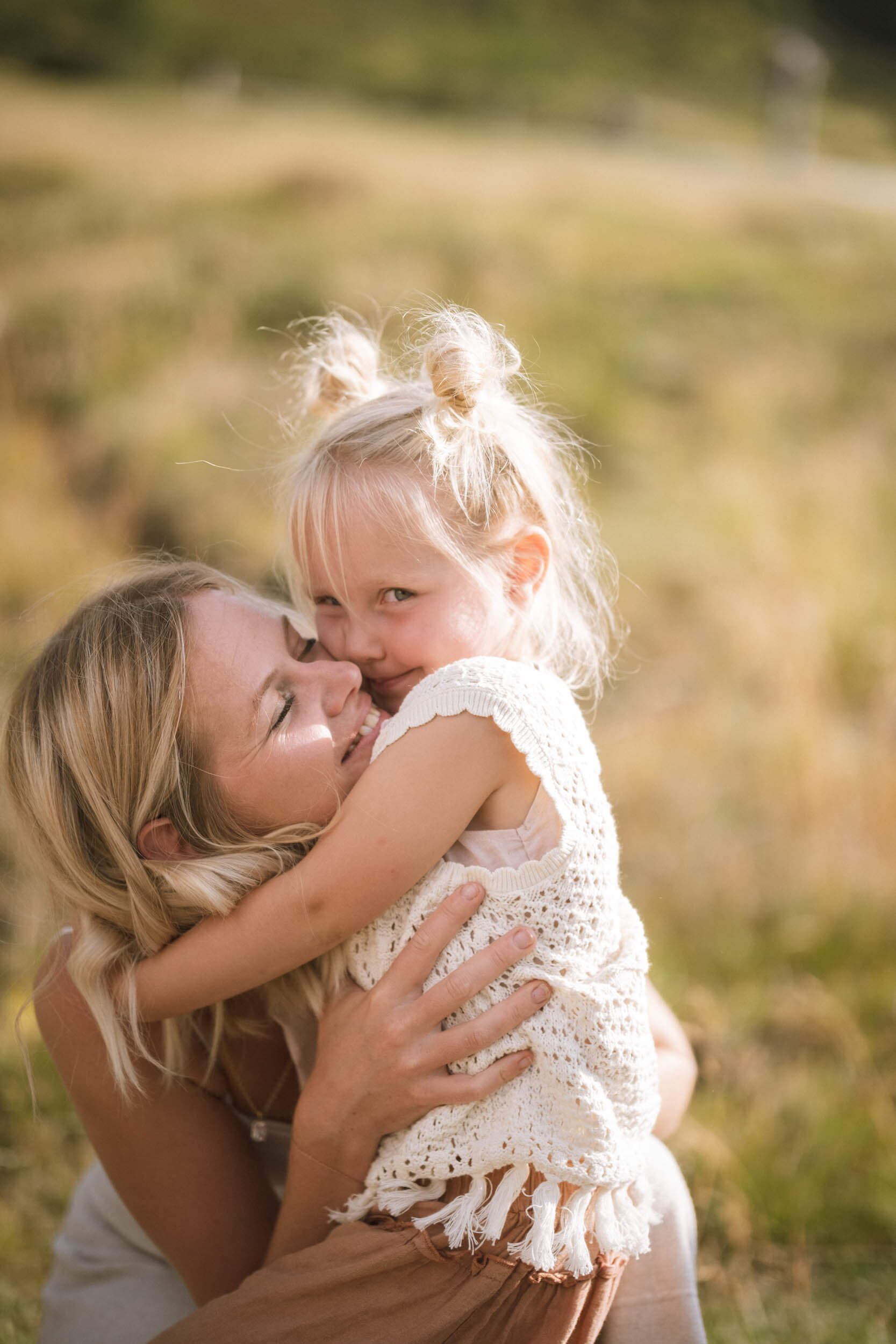 Eine lachende Frau umarmt ein kleines, lachendes Mädchen im Freien bei Sonnenuntergang.