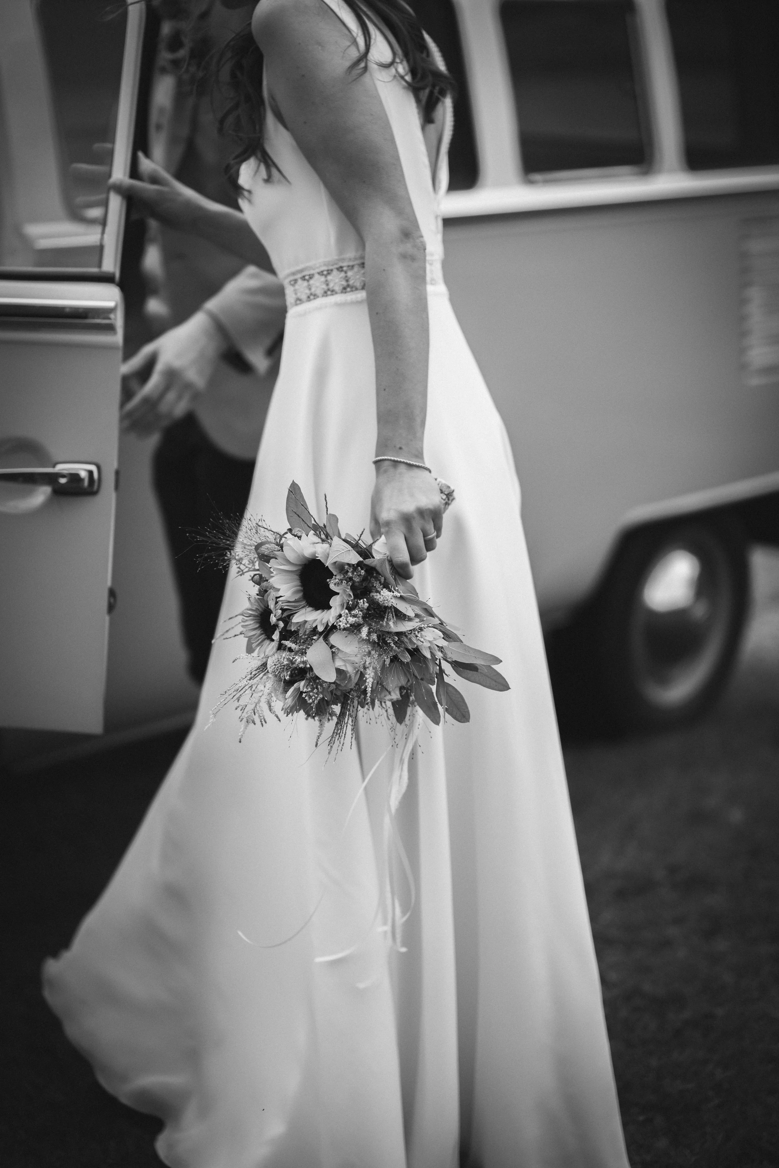 Braut in Hochzeitskleid hält einen Blumenstrauß, im Hintergrund ein Auto.