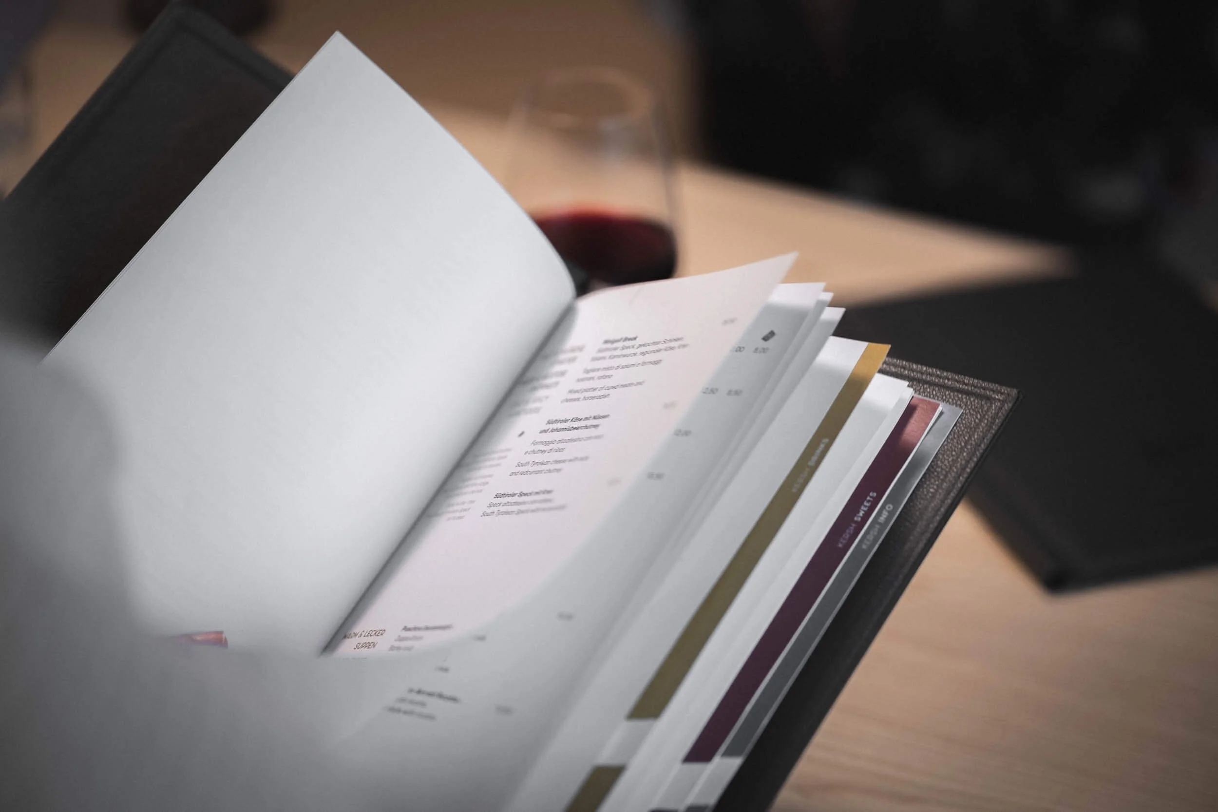 Offener Speiseplan oder Menü mit Text und Farbcodes, auf einem Tisch neben einem Glas Rotwein.