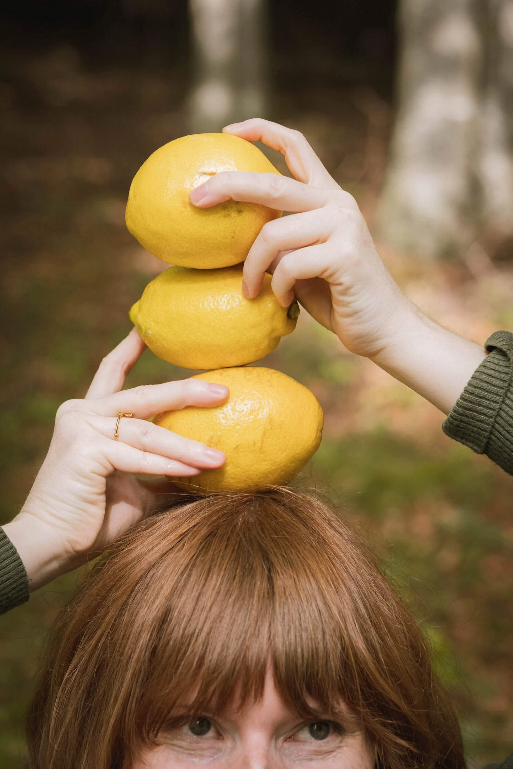Eine Person mit rotbraunem Haar hält drei Zitronen übereinander auf ihrem Kopf, im Hintergrund ein Wald.