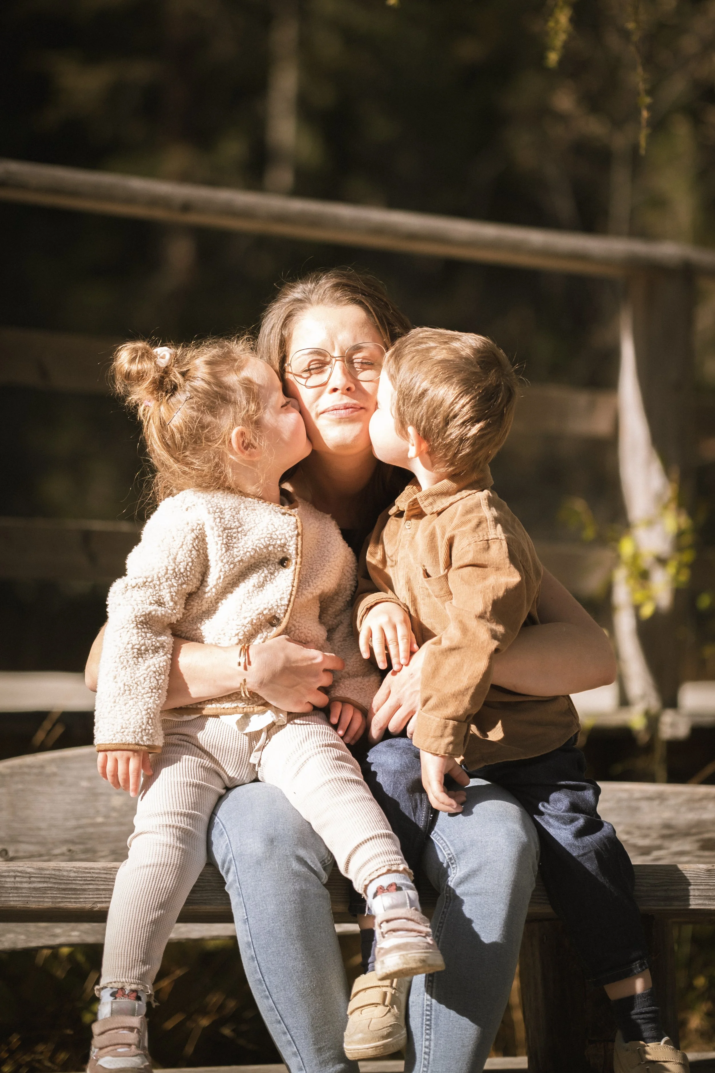 Eine Frau sitzt auf einer Bank im Freien, umgeben von Bäumen. Sie umarmt zwei Kinder, die ihr liebevoll Küsse auf die Wangen geben. Es ist sonnig und die Stimmung ist liebevoll und fröhlich.