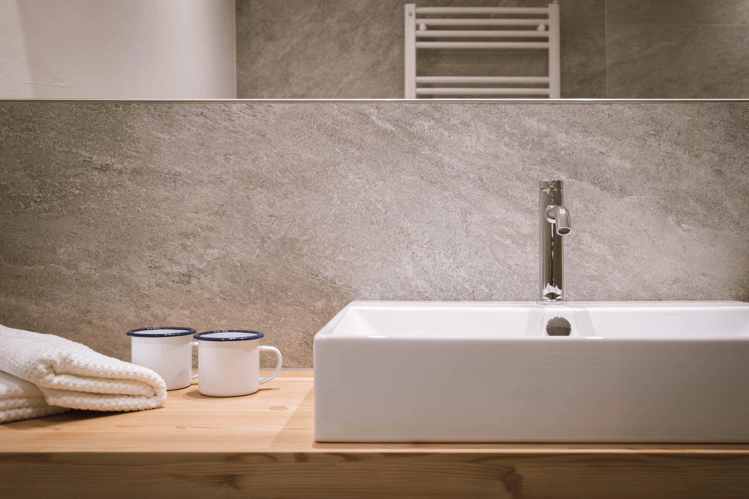 Waschbecken auf einem Holz-Waschplatz mit Handtüchern, Tassen und einem Wasserhahn, Spiegel an der Wand im Badezimmer.