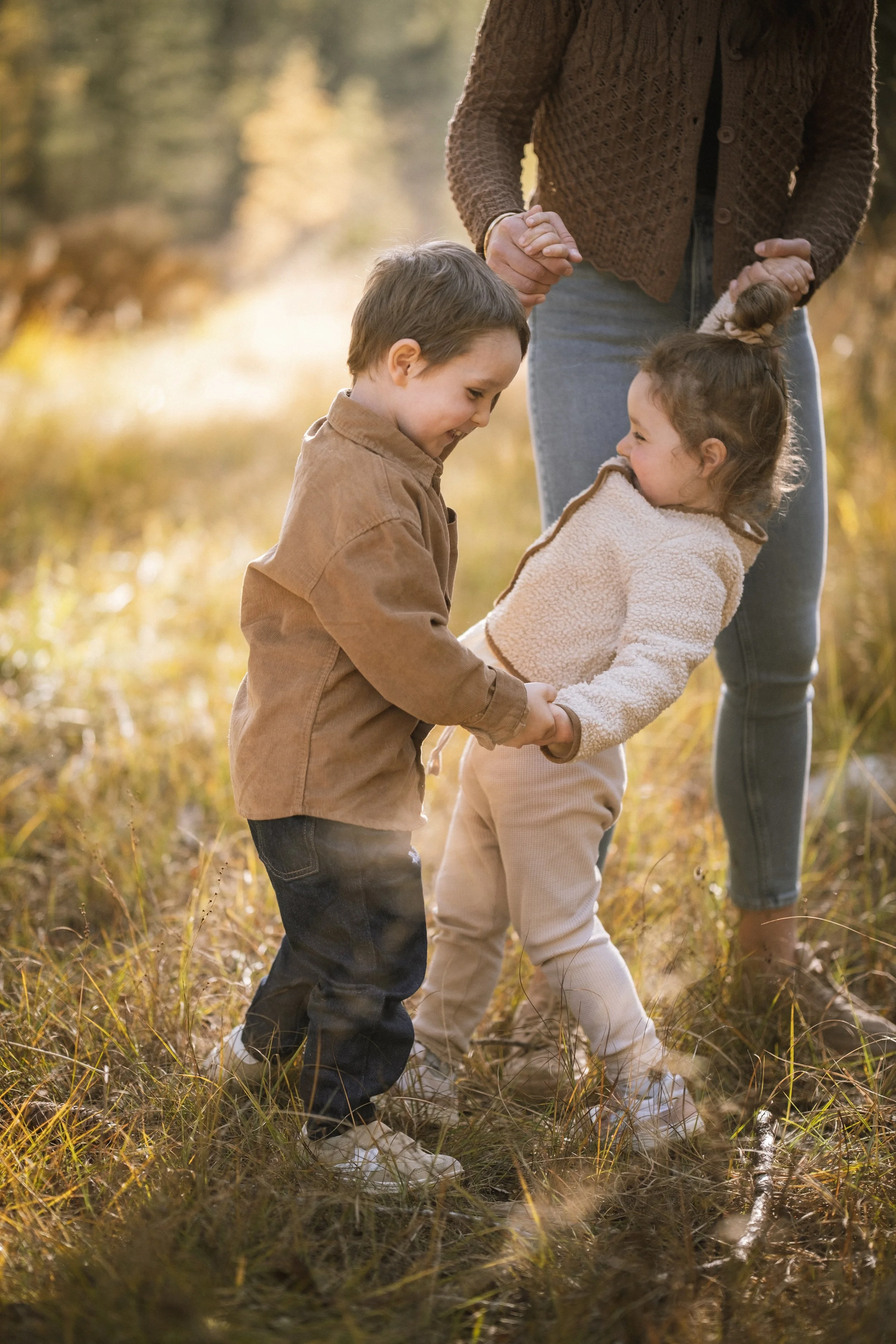 Zwei Kinder, ein Junge und ein Mädchen, spielen im Freien, während eine erwachsene Person sie dabei hält und lächelt. Es ist herbstlich, die Umgebung ist grün und die Kinder lachen und genießen die Zeit.