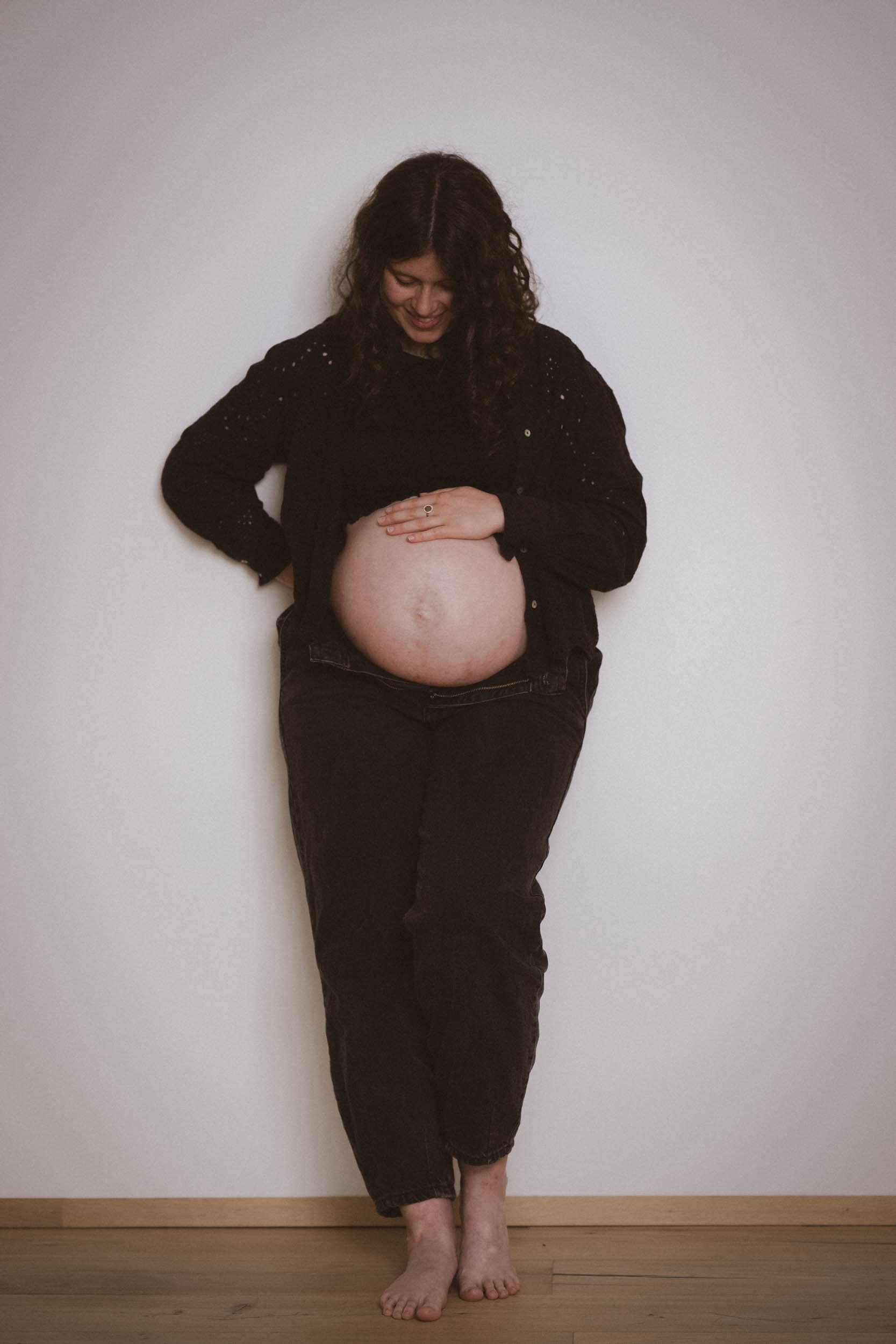 Eine schwangere Frau steht barfuß vor einer weißen Wand, lächelt und legt ihre Hand auf den Bauch, während sie nach unten schaut.