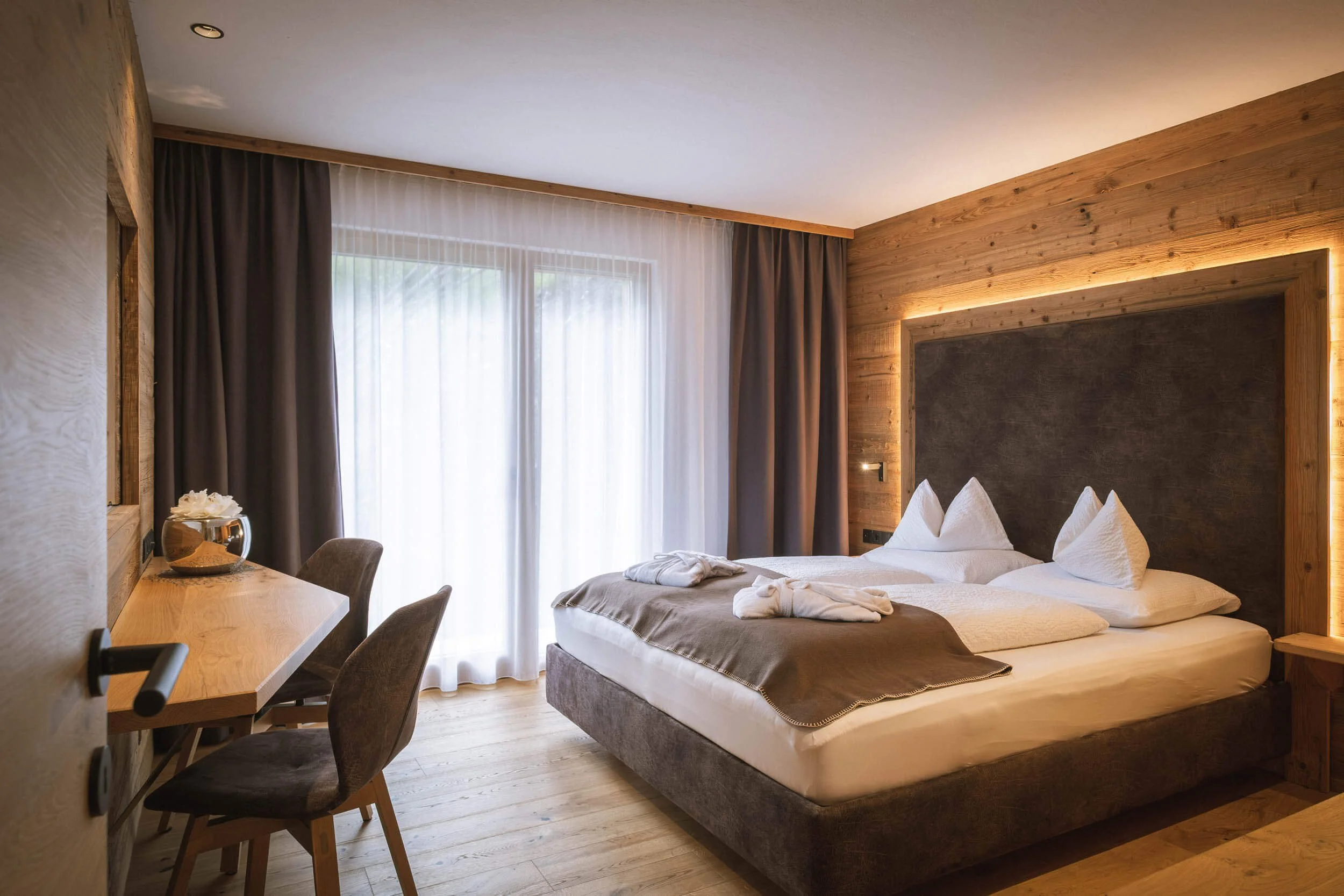 Schlafzimmer mit Doppelbett, Holz-Wandverkleidung, Nachttisch, Sonnenschutzvorhänge, Tageslicht, gedeckte und helle Farben.