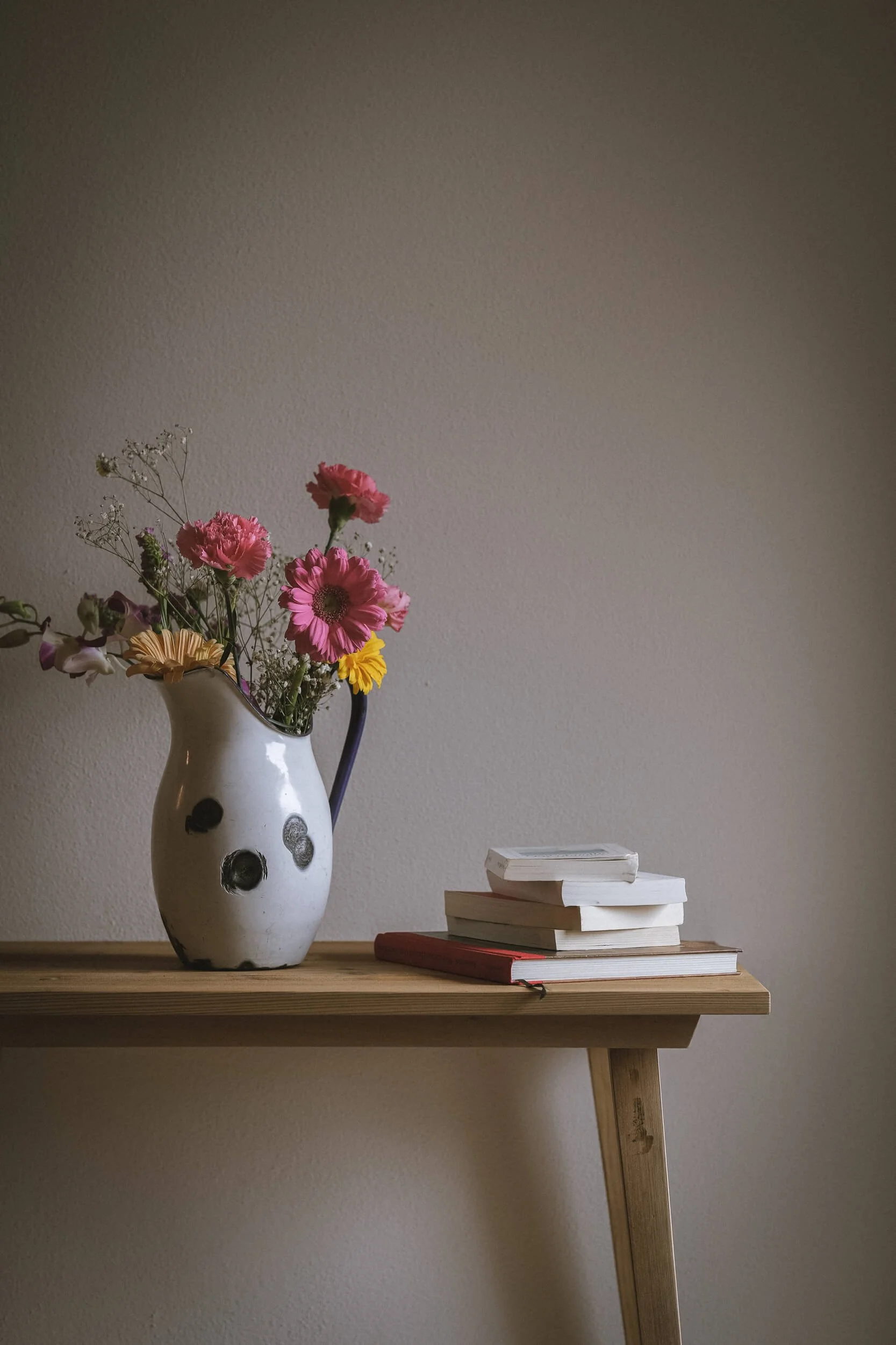 Blumen in einer weißen Vase neben einem Stapel Bücher auf einem Holztisch, gegen eine beige Wand.