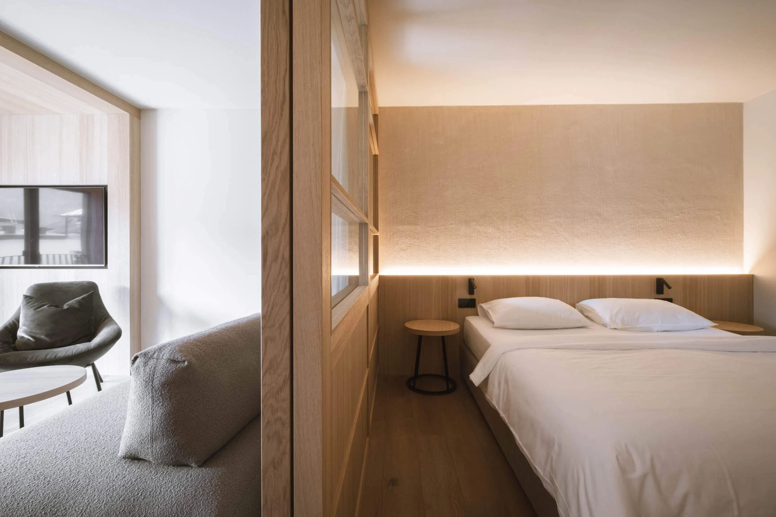 Ein Schlafzimmer mit Doppelbett, Holzverkleidung, seitlicher Nachttisch und moderner Beleuchtung, getrennt durch eine Holztrennwand von einem Wohnzimmer mit Sitzmöbeln und Fernseher.