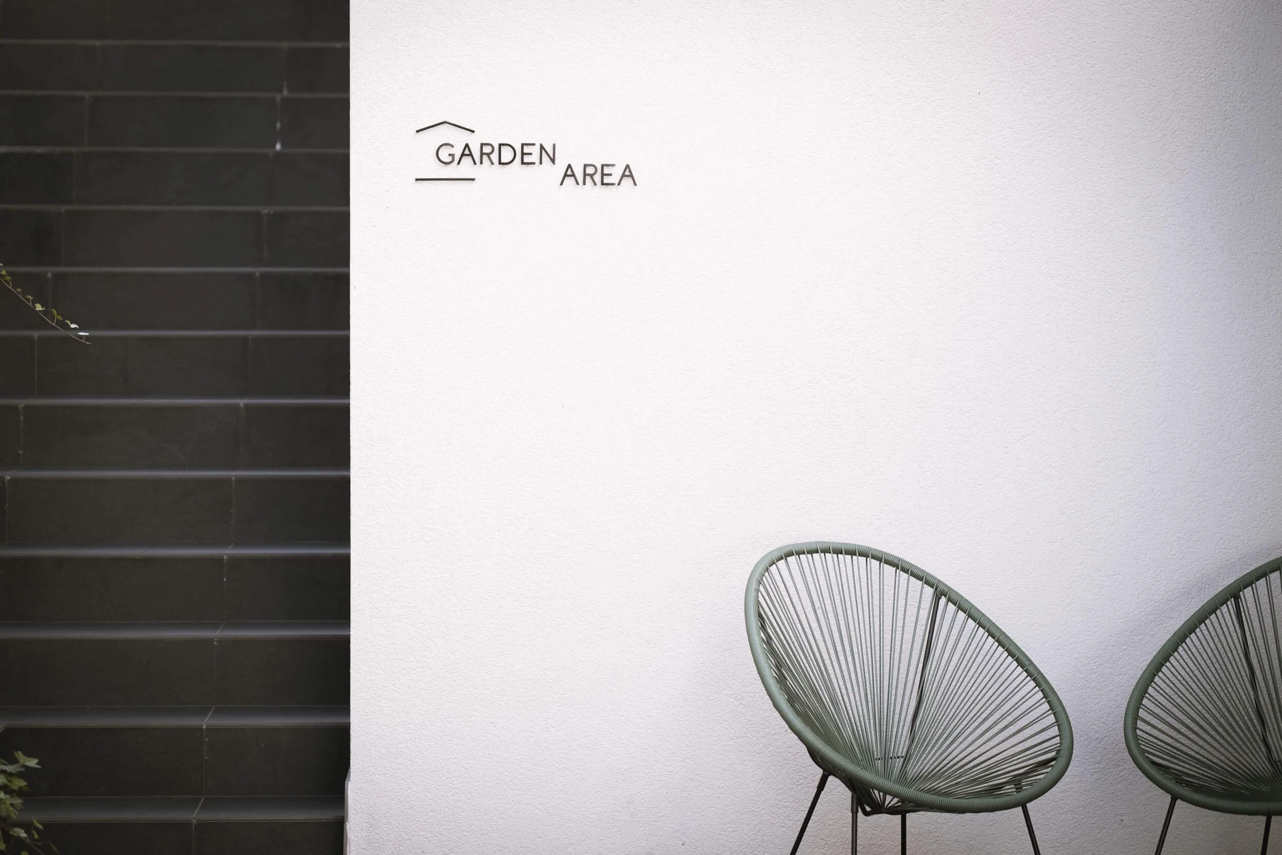 Schwarzweißes Bild eines weißen Wandteils mit der Beschriftung 'GARDEN AREA' und zwei modernen, grünen, stachelartigen Gartenstühle auf der rechten Seite neben der Wand. Auf der linken Seite befinden sich schwarze Fliesen.