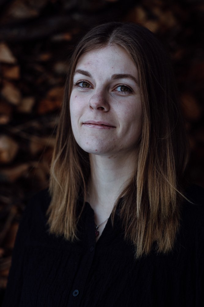Porträt einer jungen Frau mit braunen Haaren vor einem Hintergrund aus Holzscheiten.