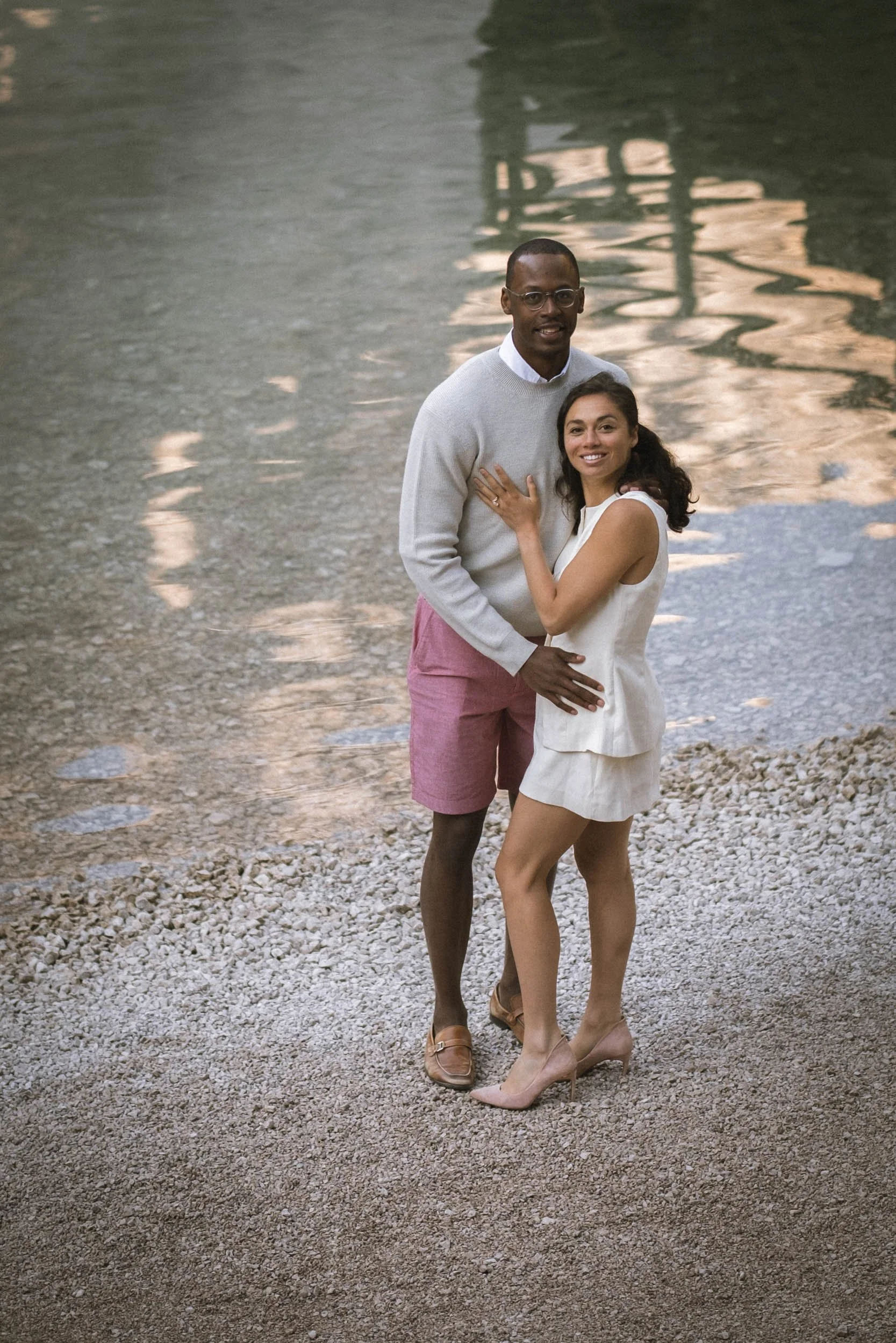 Ein Paar steht am Flussufer, die Frau umarmt den Mann. Sie lachen und wirken glücklich, im Hintergrund Wasser und Felsen.