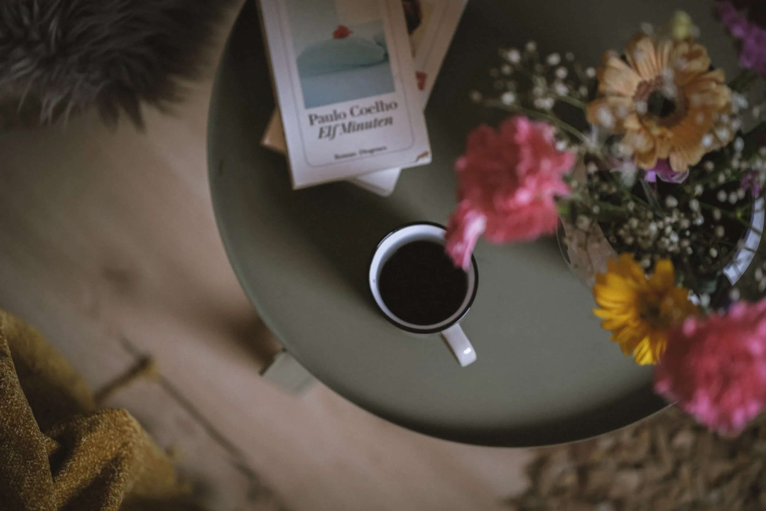 Eine graue Tischplatte mit einer Kaffeetasse, einer Blumenvasen mit bunten Blumen und einer Packung Paojo Coelho El Jardin.