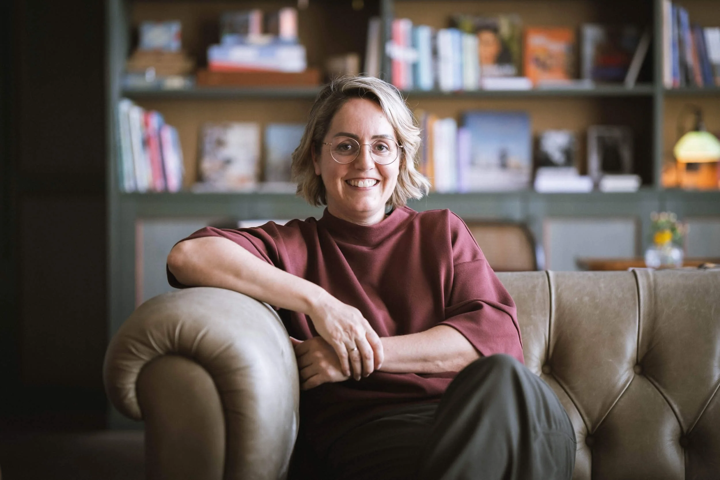 Eine lächelnde Frau mit Brille sitzt auf einem braunen Ledersofa in einem Raum mit Buchregalen im Hintergrund.