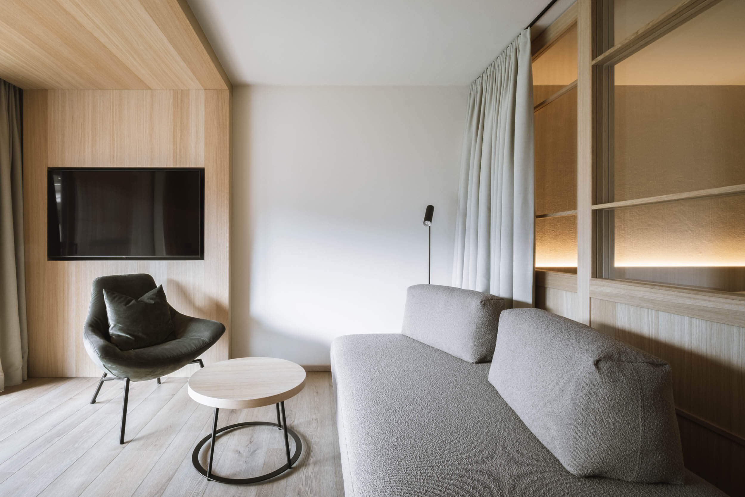 Moderner Wohnzimmerbereich mit grauem Sofa, schwarzen Sessel, kleinem runden Holztisch, Wandmontiertem Fernseher, weißen Vorhängen und Holzverkleidungen.