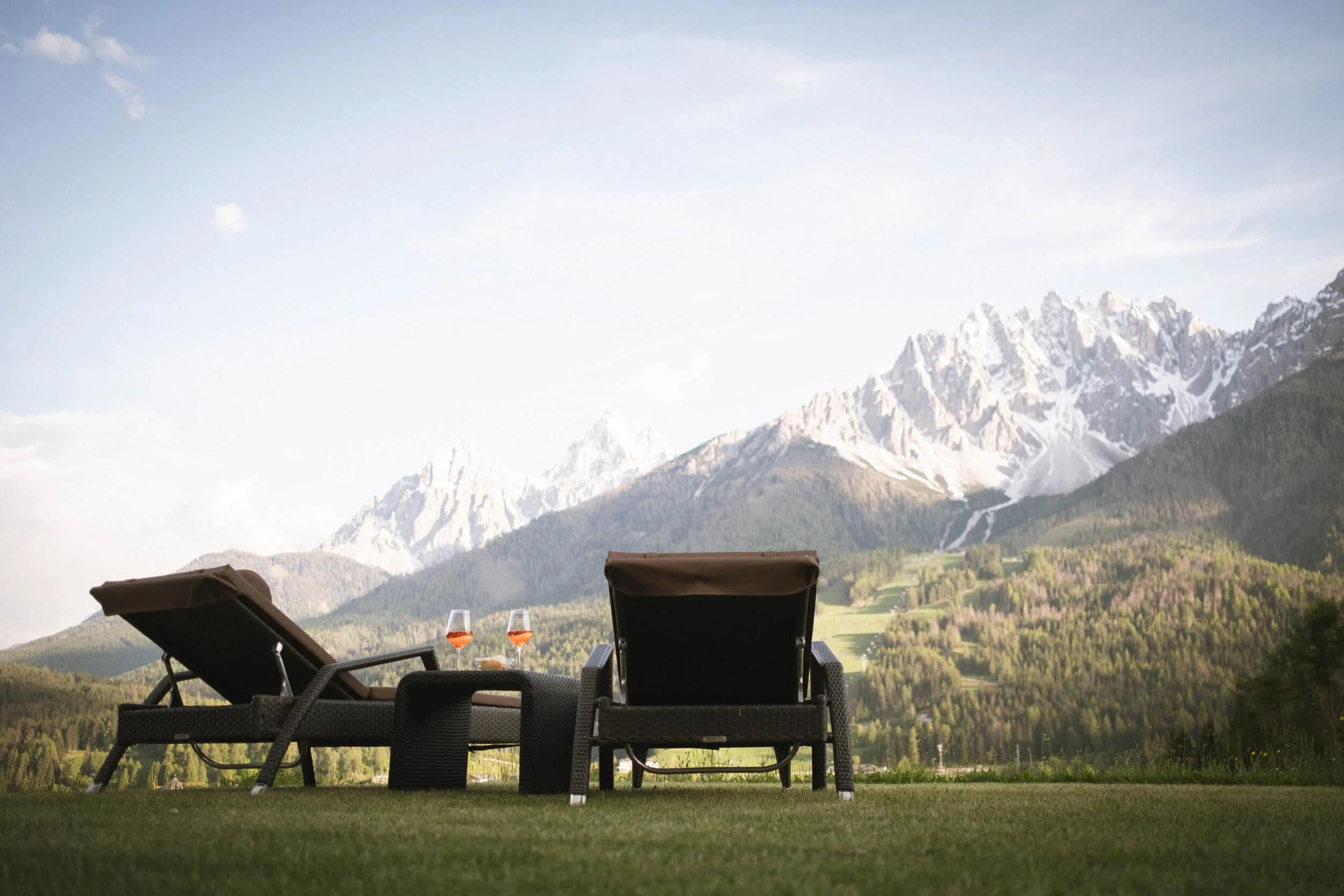Zwei Liegestühle auf einer Wiese mit zwei Gläsern Wein vor einer Bergkulisse, schneebedeckte Berge im Hintergrund.