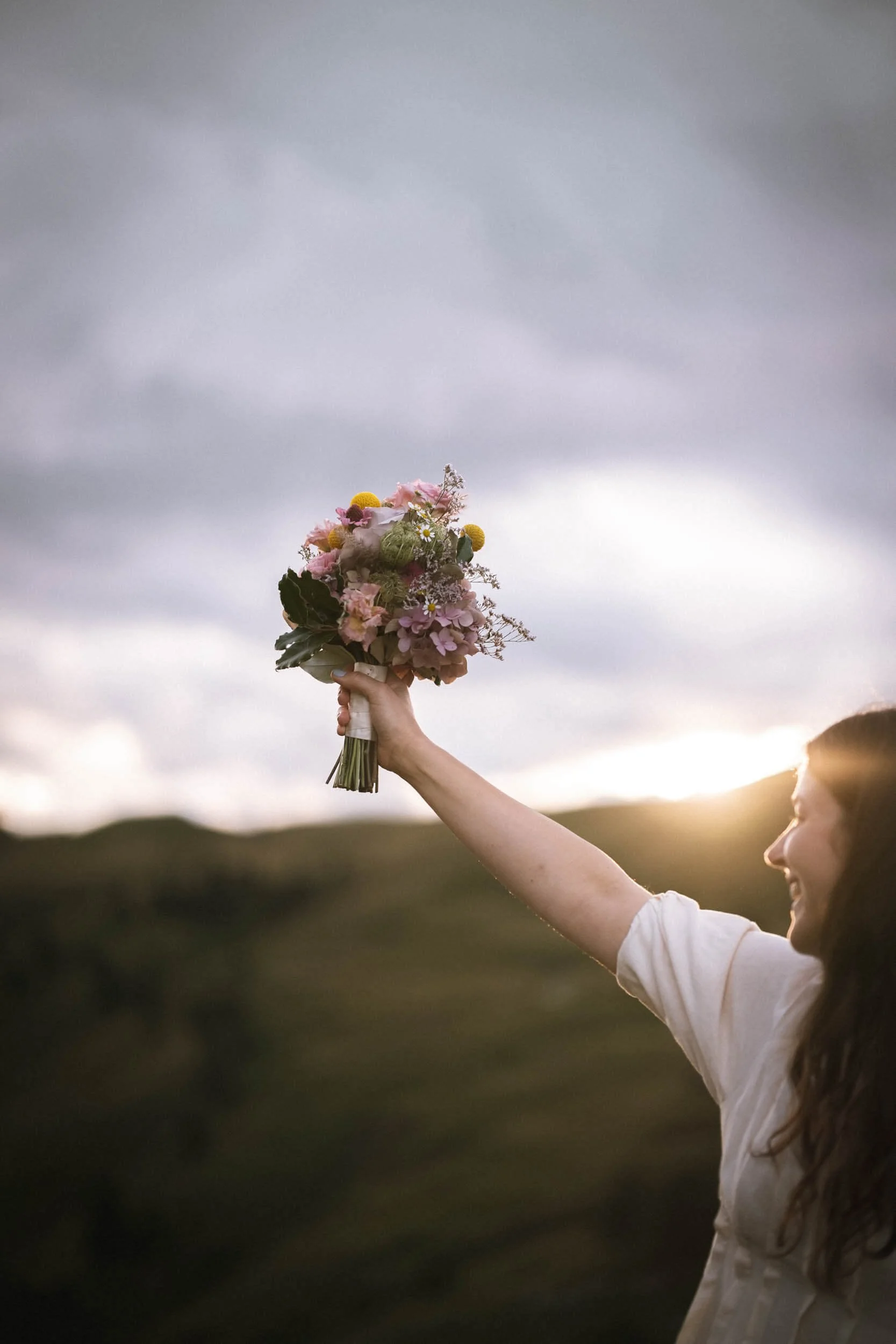 Frau hält einen bunten Blumenstrauß im Sonnenuntergang in der Hand hoch, im Hintergrund eine bewölkte Landschaft.