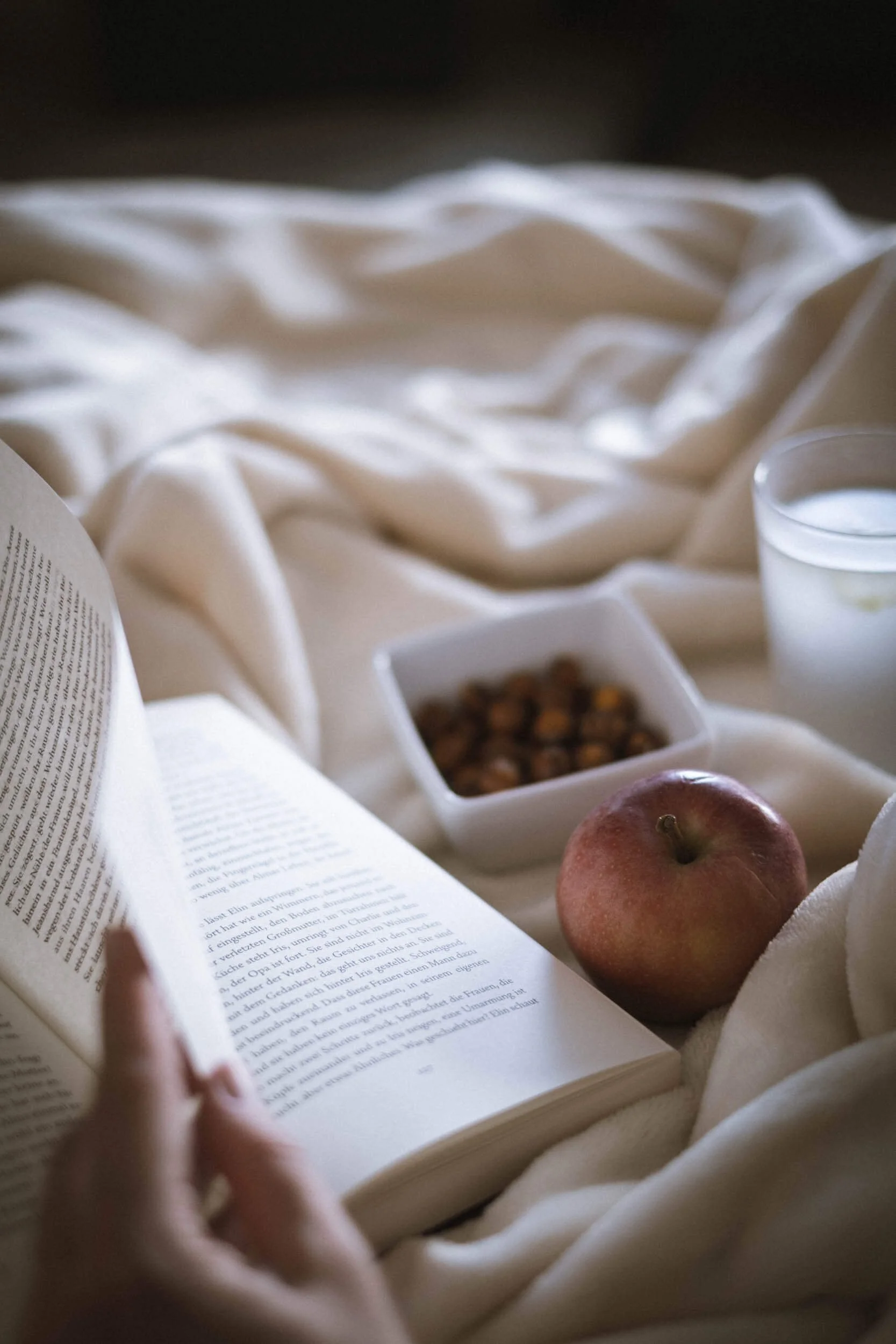 Eine Person liest ein Buch, umgeben von einem Apfel, einer Schüssel mit Nüssen, einem Glas Wasser und einem Glas Milch auf weißem, weichem Stoff. Das Bild ist in warmem Licht gehalten.