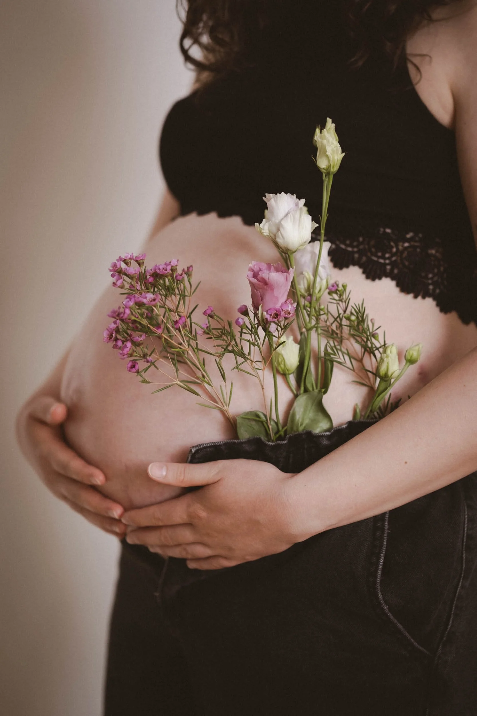 Schwangere Frau hält Blumen vor ihren Bauch, die aus ihrem Bauch herauswachsen, und trägt ein schwarzes Oberteil und dunkle Jeans.