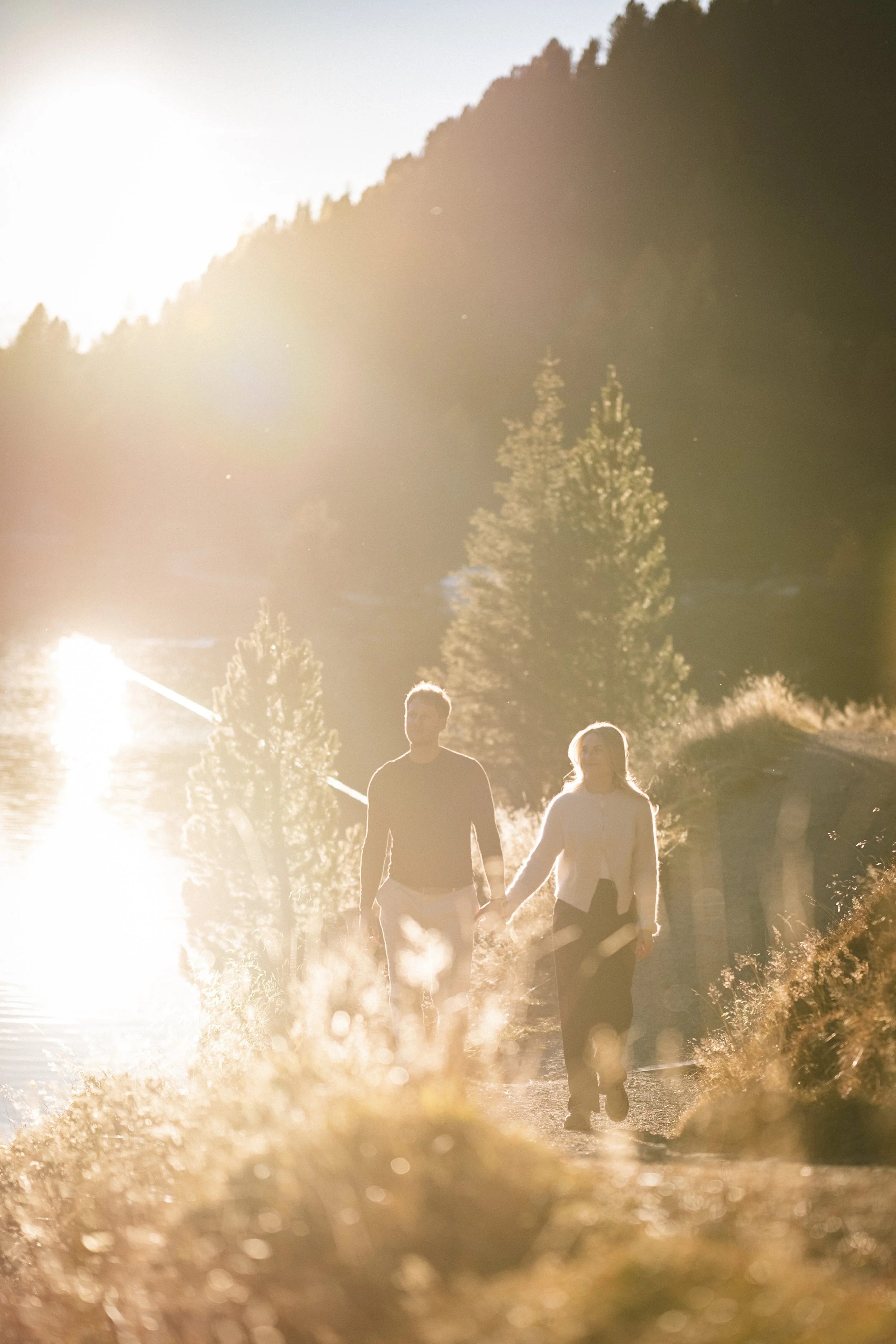 Ein Mann und eine Frau gehen Hand in Hand an einem See bei Sonnenuntergang, umgeben von Bergen und Bäumen.