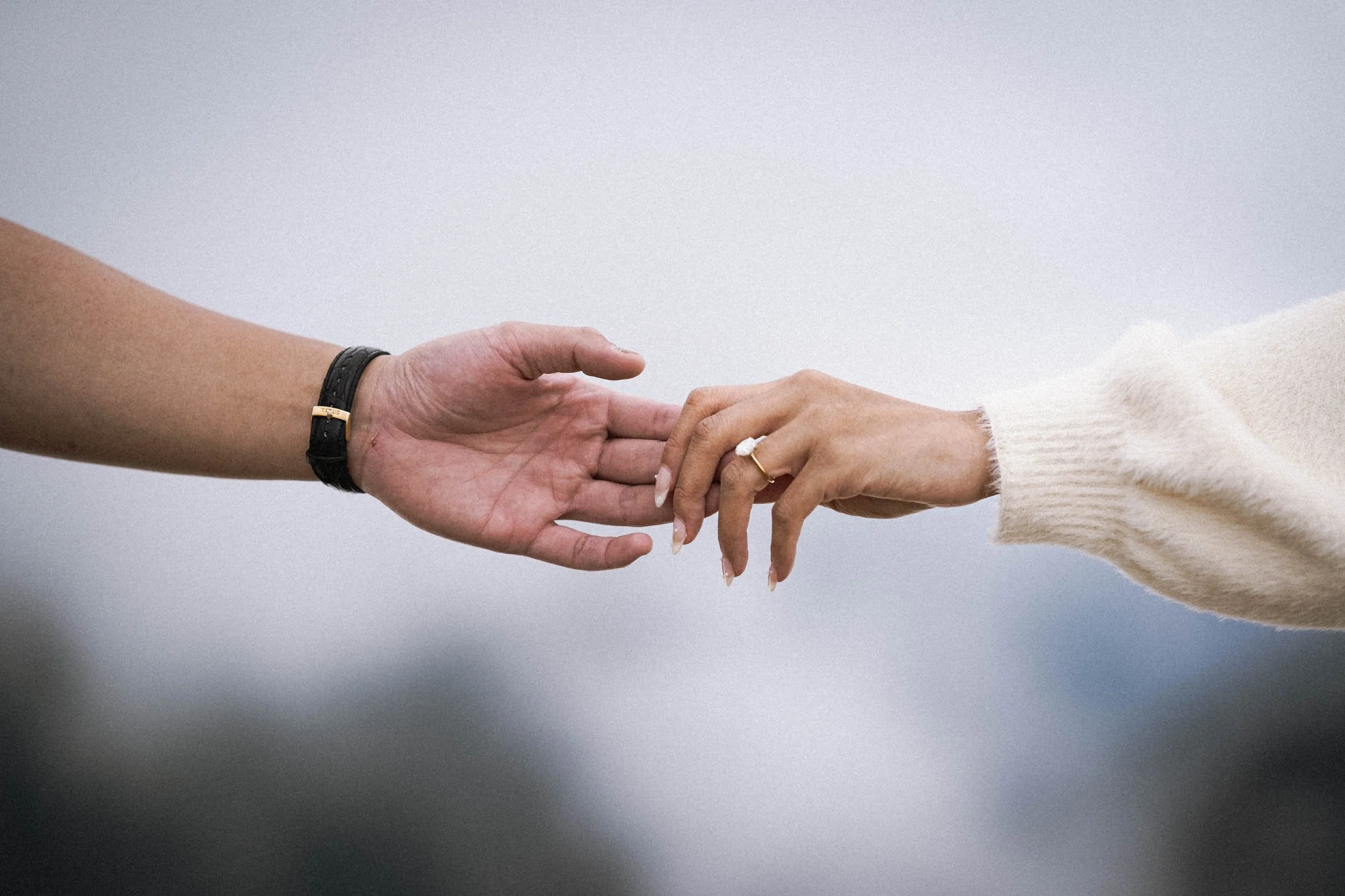 Zwei Hände, eine männlich mit dunkler Haut, die andere weiblich mit heller Haut, greifen sich an den Fingern an einem neutralen Hintergrund.