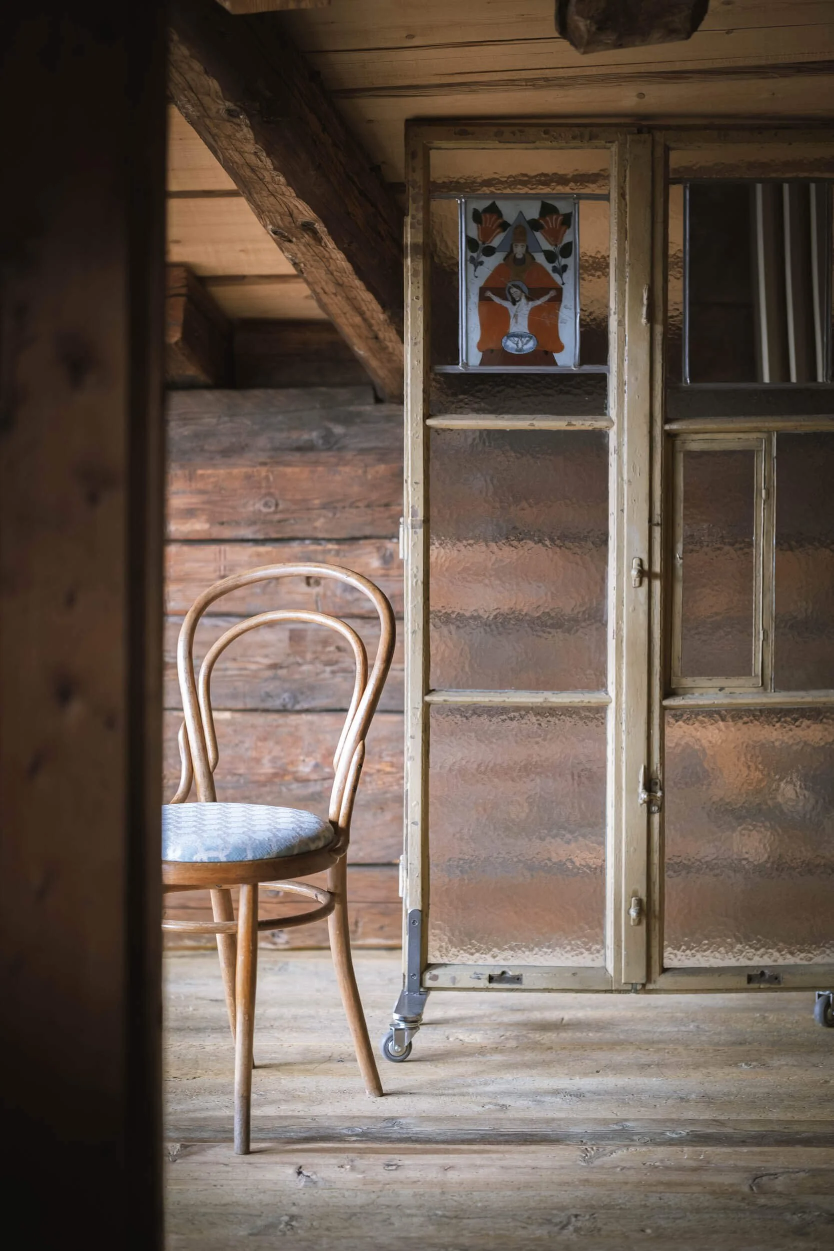 Ein Stuhl aus Holz mit einem gepolsterten Sitz in einem Raum mit Holzboden und Holzdecke, durch eine Glas- und Holzschiebtür sichtbar.