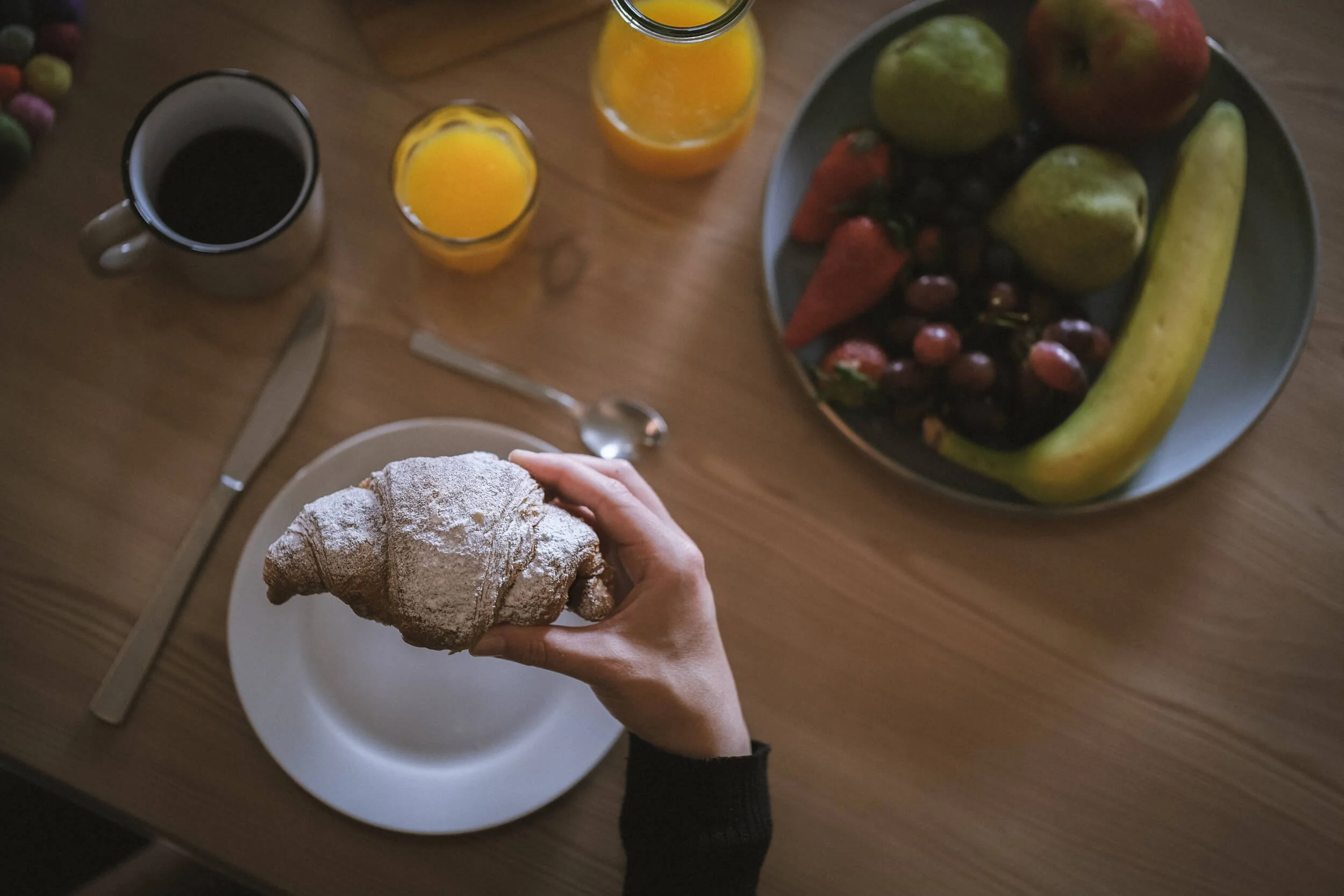 Hand hält ein mit Puderzucker bestäubtes Croissant auf einem weißen Teller. Auf dem Tisch sind ein Messer, eine Gabel, zwei Gläser mit Orangensaft, eine Tasse Kaffee und eine große Schüssel mit frischem Obst (Äpfel, Birnen, Trauben, Karotten, Banane)