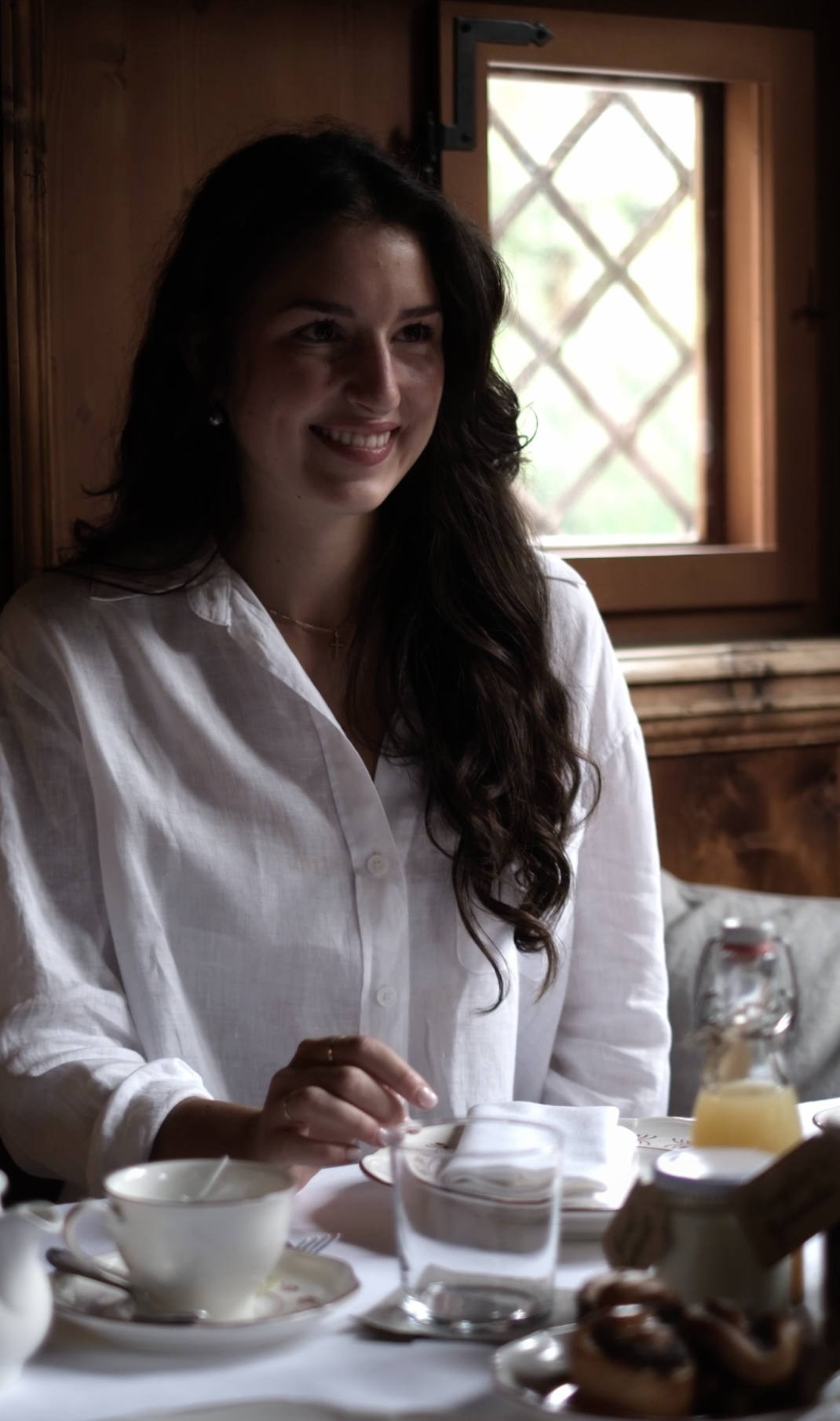 Eine lachende junge Frau mit dunklen, lockigen Haaren und weißem Hemd sitzt an einem Frühstückstisch in einem rustikalen Raum mit Holzwänden und einem offenen Fenster im Hintergrund.