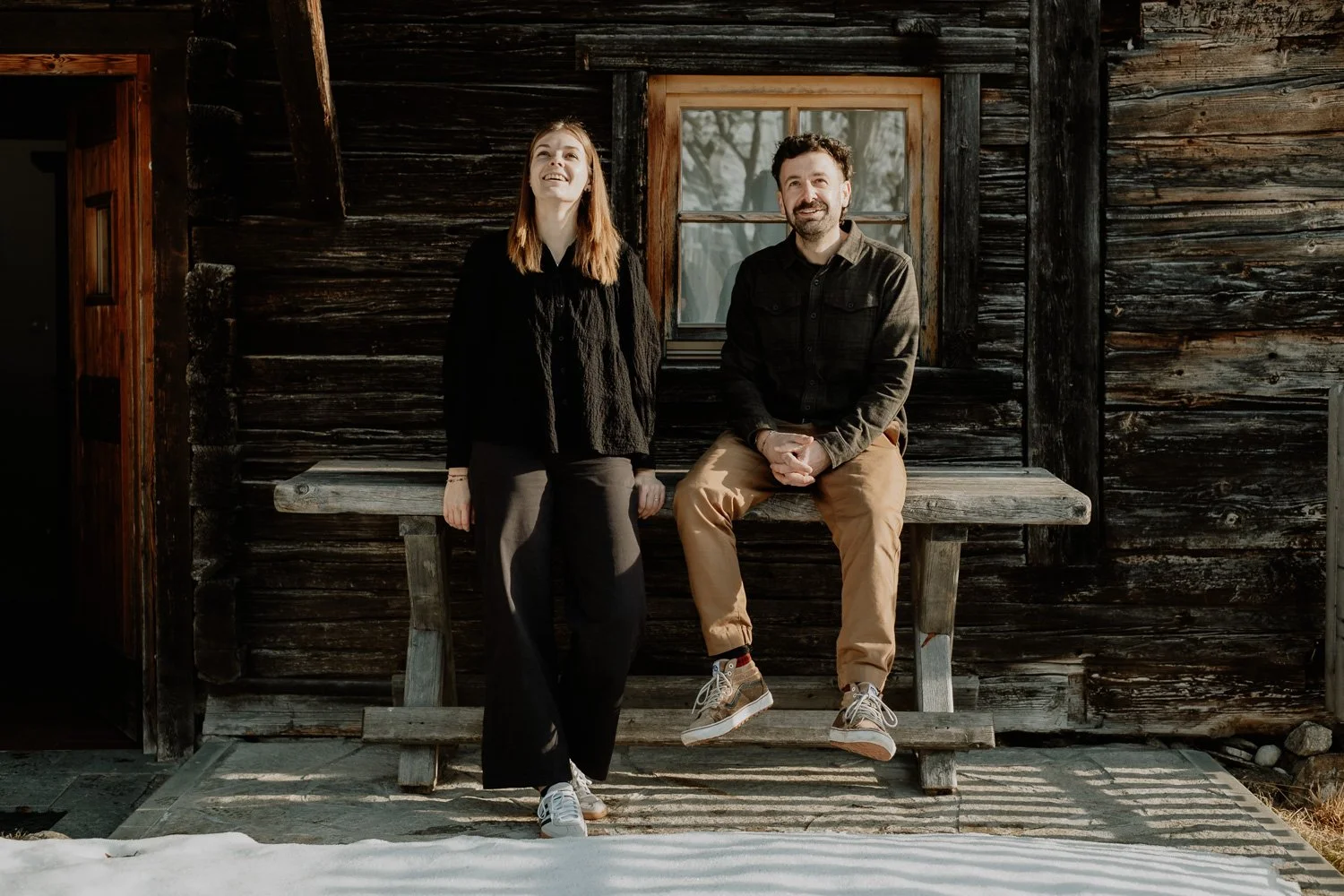 Ein Mann und eine Frau posieren vor einer alten Holzhütte, die von Sonnenlicht beleuchtet wird.